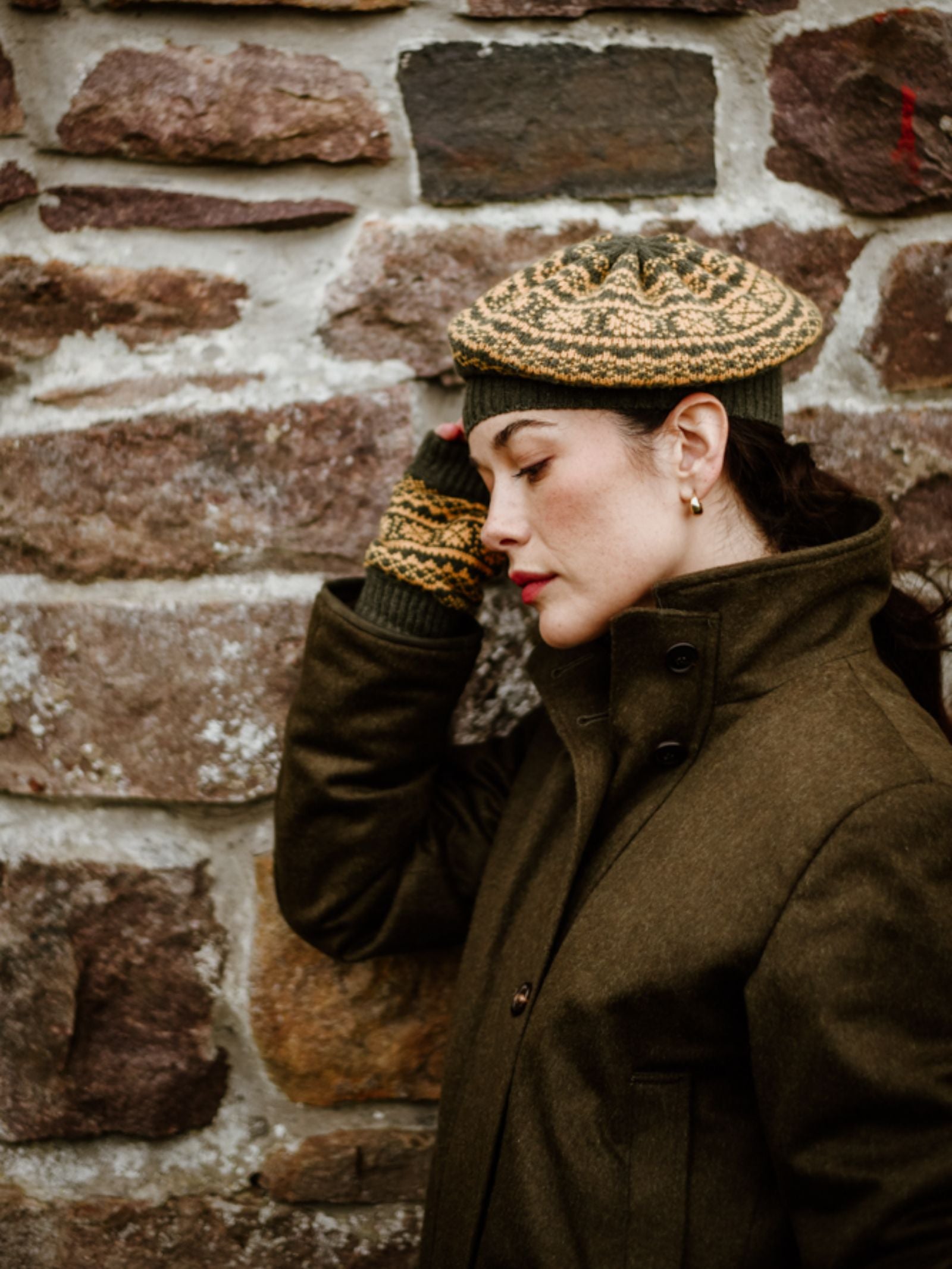 A woman in an olive coat, Campbell's of Beauly 2 Colour Fairisle Beret, and Scottish knitwear fingerless gloves leans against a stone wall, touching her head and looking down thoughtfully.
