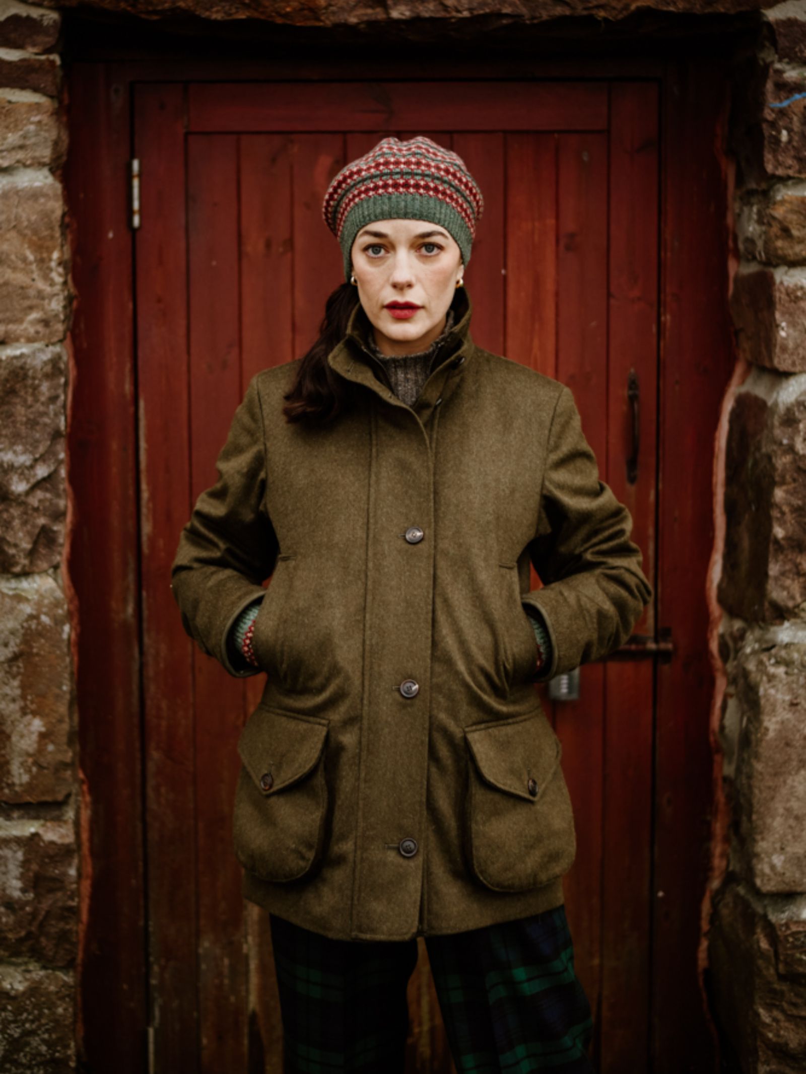 A woman stands before a rustic red door in a stone wall, wearing Campbell's of Beauly's Loden Green Field Coat made from Austrian loden fabric, along with plaid pants and a knitted hat, hands in pockets, facing the camera.