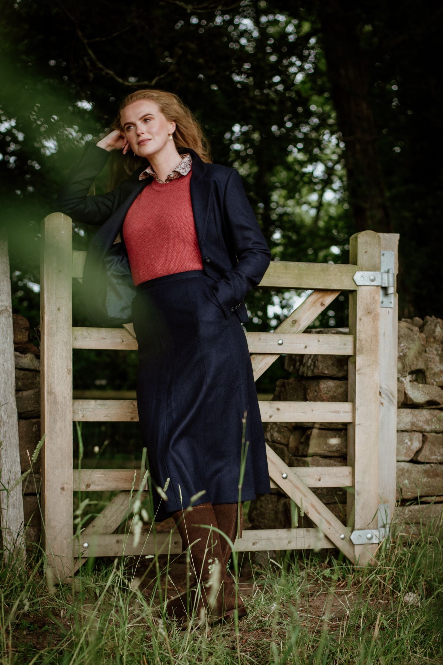 A woman with long hair stands outdoors by a wooden gate, wearing a dark coat, red top, and the Campbell's of Beauly Panel Skirt. She looks thoughtful, resting her head on one hand amid lush greenery and trees.