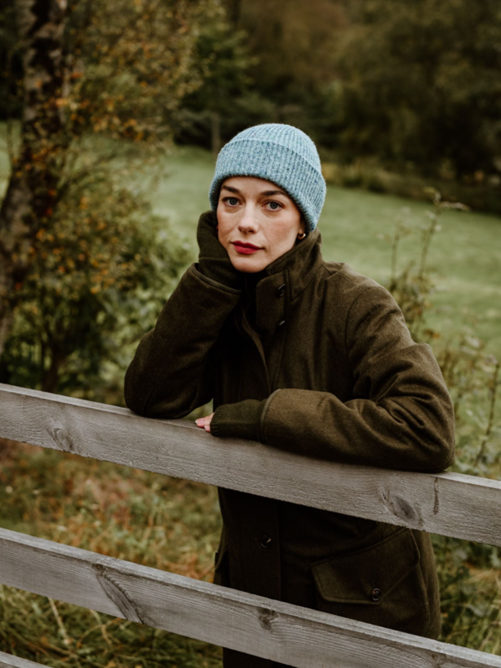 A woman in a Campbell's of Beauly Loden Green Field Coat and a light blue knit beanie leans on a wooden fence, resting her chin on her hand, with grassy woods behind her.