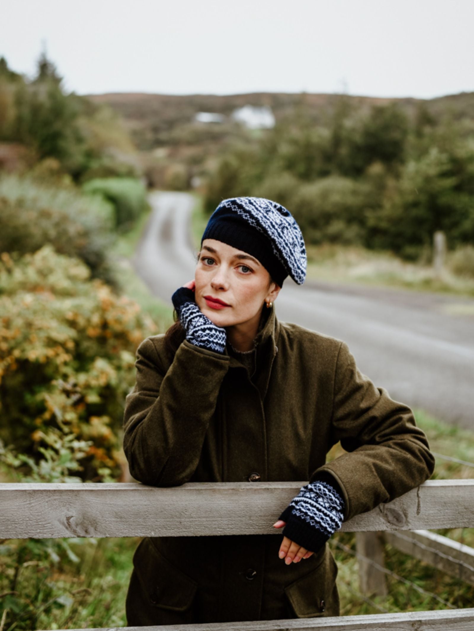 A woman in a green coat wears the Campbell's of Beauly 2 Colour Fairisle Beret with matching fingerless gloves, leaning on a wooden fence by a rural road amid greenery, looking thoughtfully at the camera.