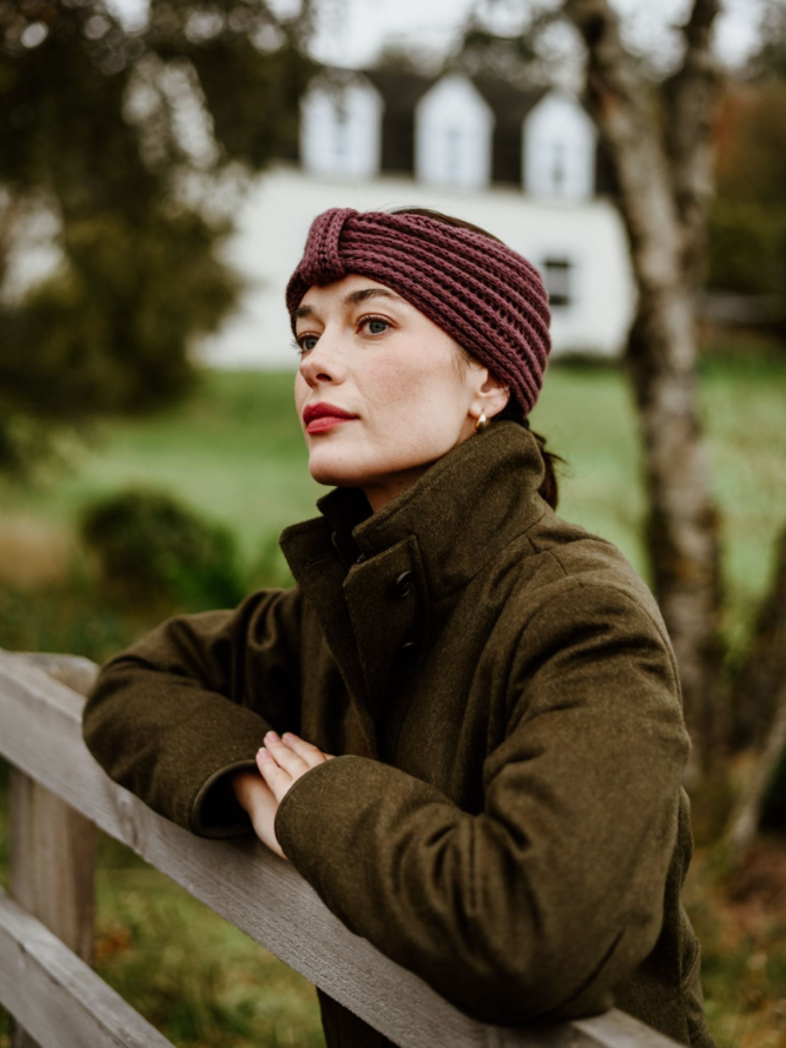 A woman in a dark coat and a Campbell's of Beauly Cashmere Ear Warmer leans on a wooden fence, gazing thoughtfully into the distance with a blurred house and greenery behind her.