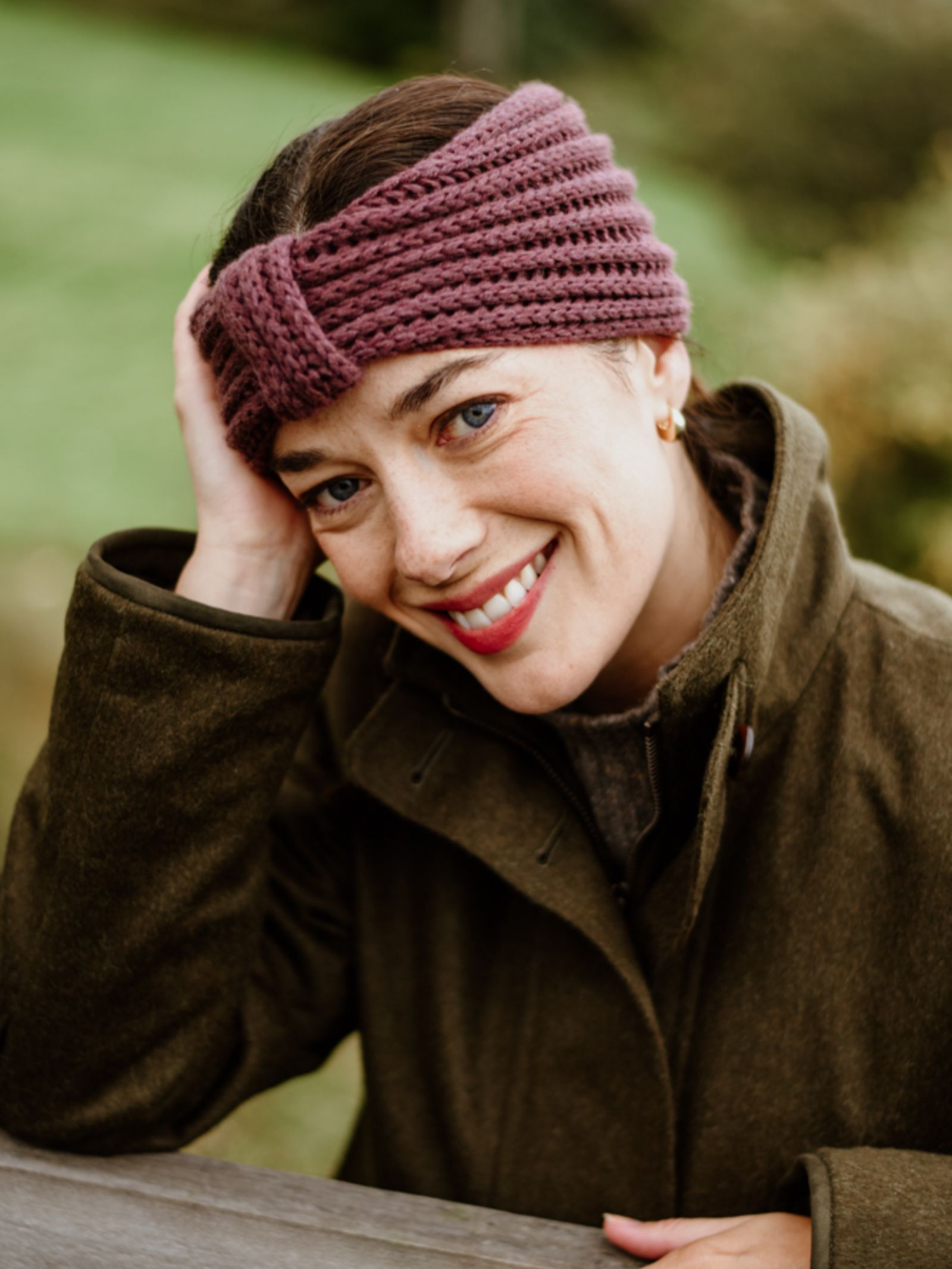 A woman outdoors wears the Campbell's of Beauly Cashmere Ear Warmer in purple with an olive green coat, smiling warmly against a backdrop of greenery—a cozy winter accessory for chilly days.