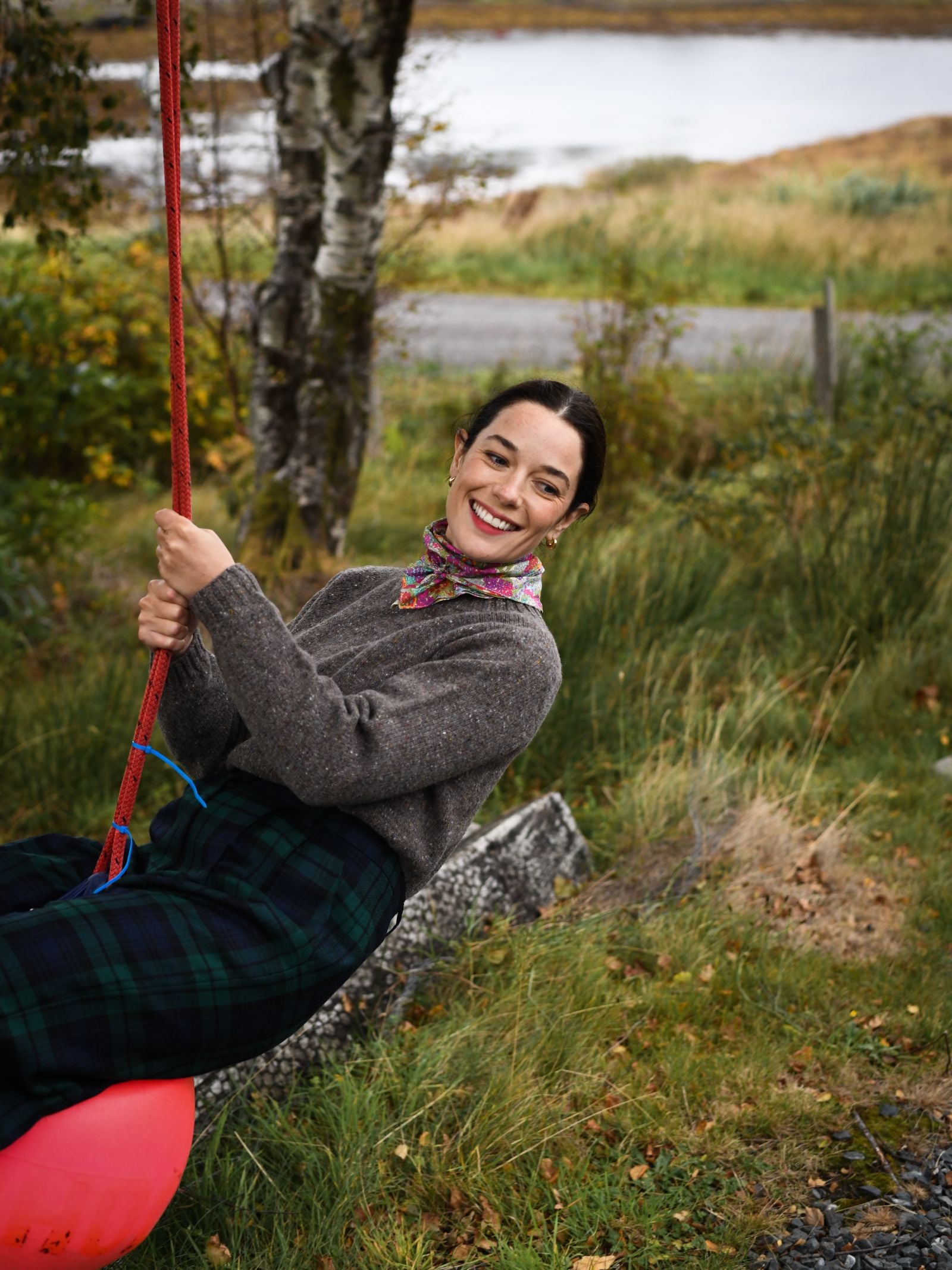 A smiling woman in a grey sweater and plaid skirt swings on a red rope swing outdoors, wearing the Campbell's of Beauly Square Headscarf as a stylish accessory. Trees, grass, and a river form the scenic background.