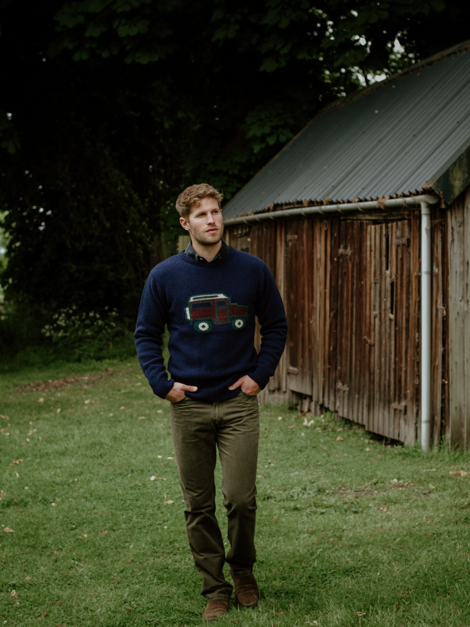 A man with light brown hair wears the Campbell's of Beauly Unisex Land Rover Defender Jumper in navy and olive green pants as he walks on grass near a rustic wooden shed, surrounded by trees and lush greenery.
