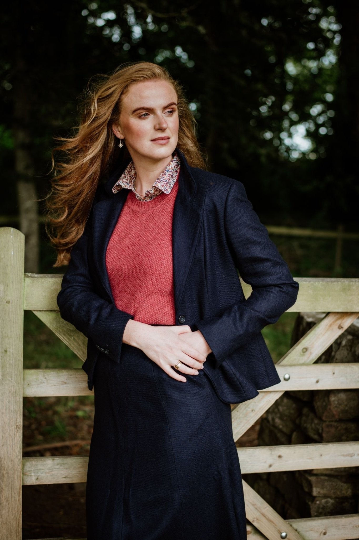 A woman with long, wavy auburn hair stands outdoors by a wooden gate, wearing Campbell's of Beauly's Panel Skirt with a navy blazer and red sweater. Green trees and a fence are visible in the background.