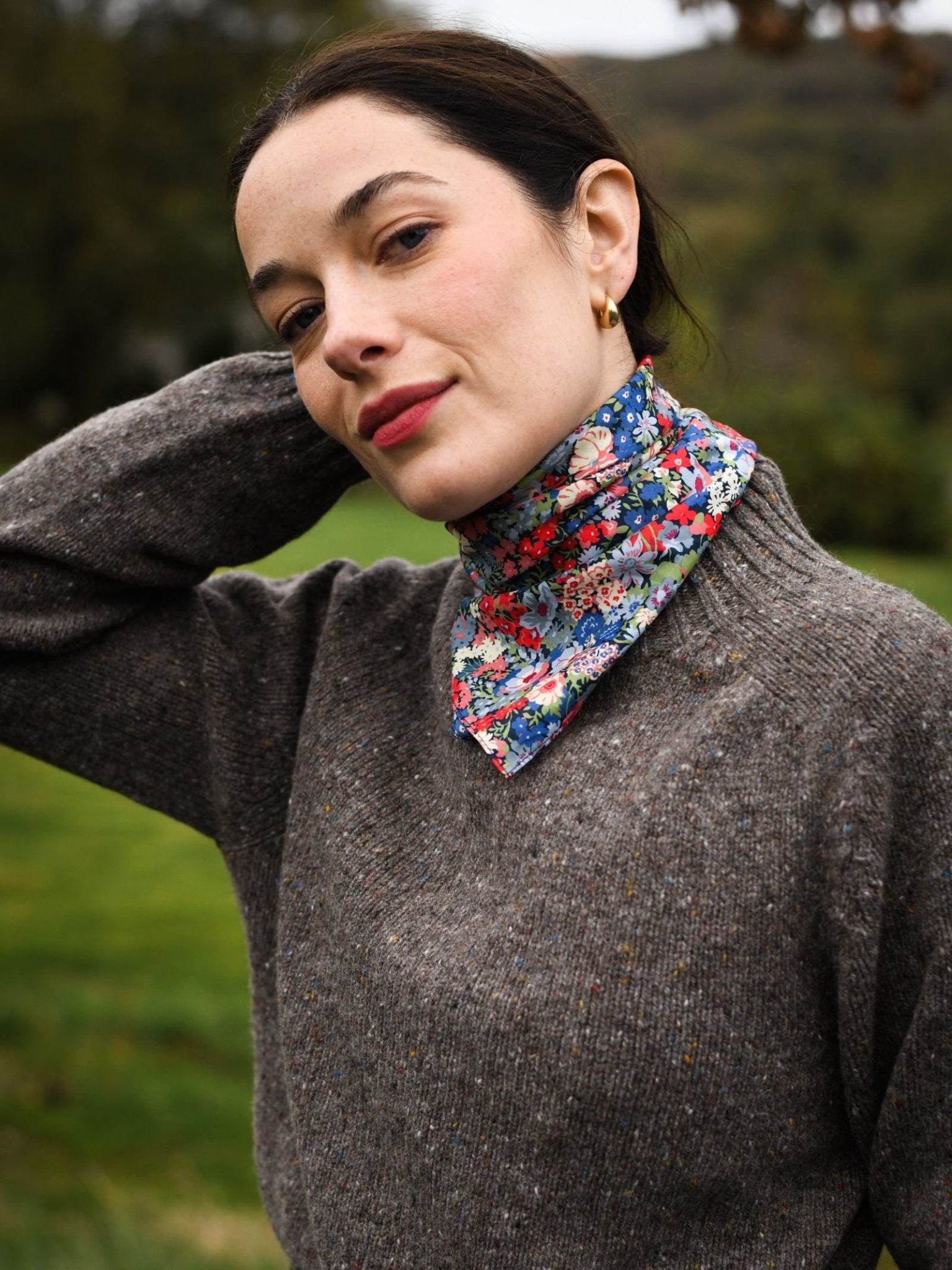 A woman with fair skin and dark hair, wearing a grey sweater and a vibrant Campbell's of Beauly Square Headscarf, stands outdoors on blurred grass, one hand behind her head, looking at the camera.