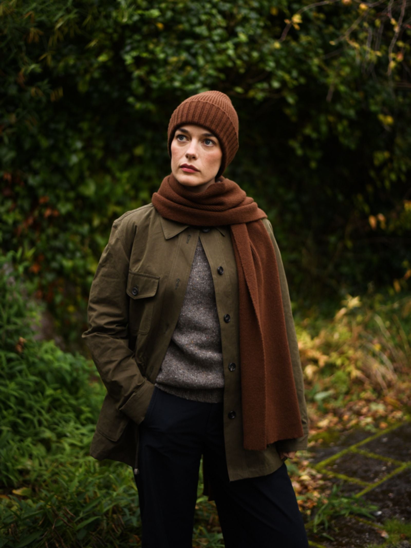 Wearing Campbell's of Beauly Rib Hat with a matching scarf, olive green jacket, and gray sweater, a person stands outdoors among green foliage, looking off to the side—showcasing classic winter style.