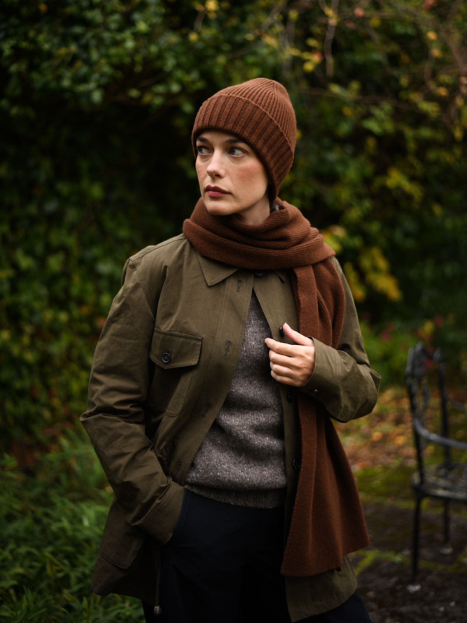 Wearing the Campbell's of Beauly Rib Hat, a brown scarf, olive green jacket, and grey sweater, this person stands in a lush garden with one hand in their pocket—a perfect winter essential look.