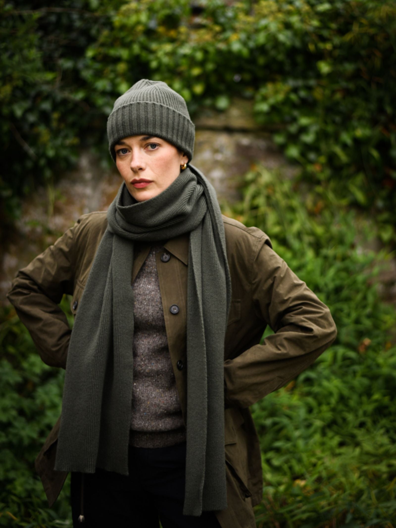 A person in a green Campbell's of Beauly Rib Hat, matching scarf, brown jacket, and gray sweater stands outdoors before leafy greenery, gazing at the camera with a serious look—showcasing this winter essential beanie.