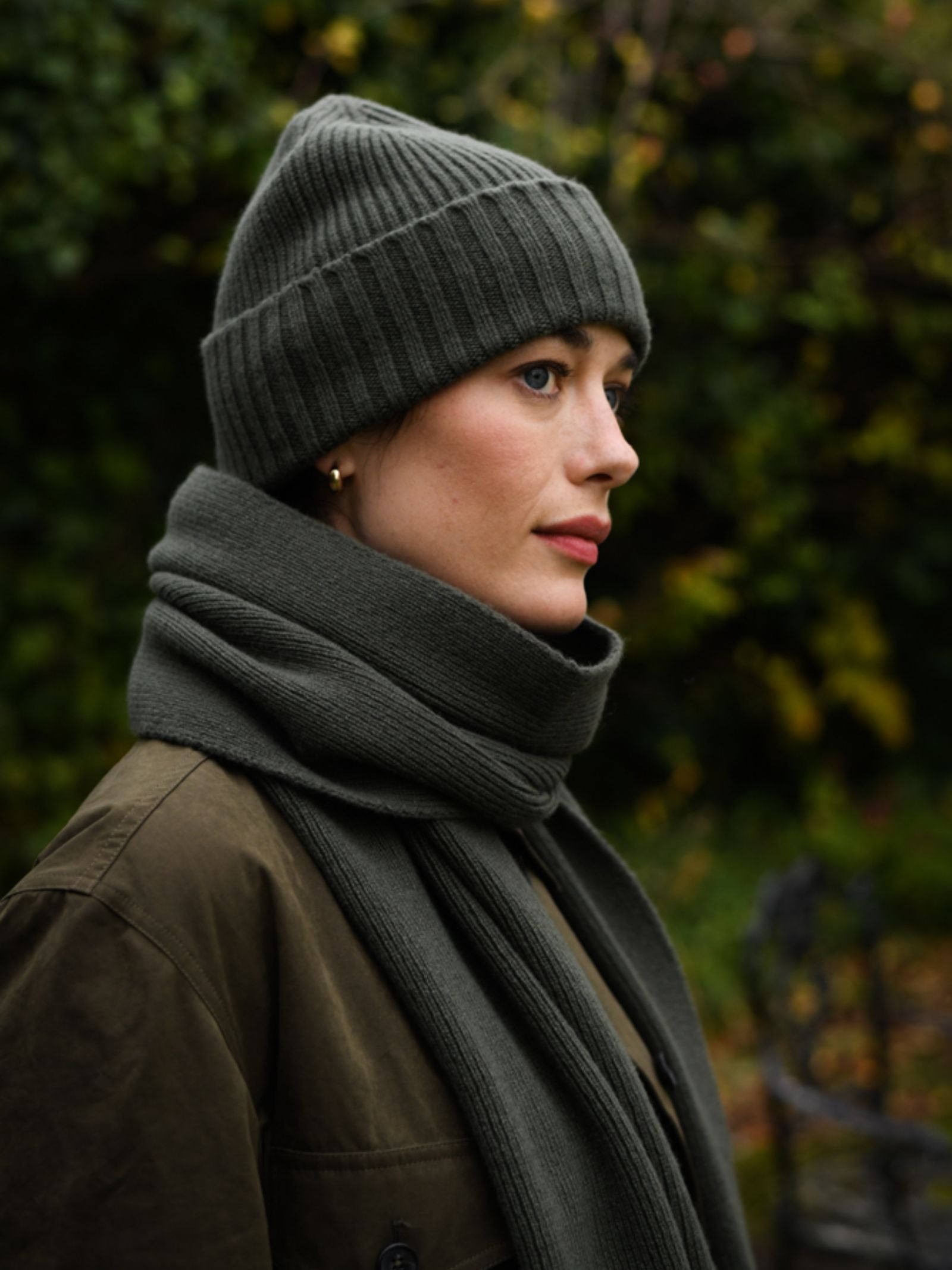 A woman stands outdoors against blurred green foliage, wearing a Campbell's of Beauly Rib Hat in dark green 100% wool with a matching scarf and brown coat—a true winter essential.