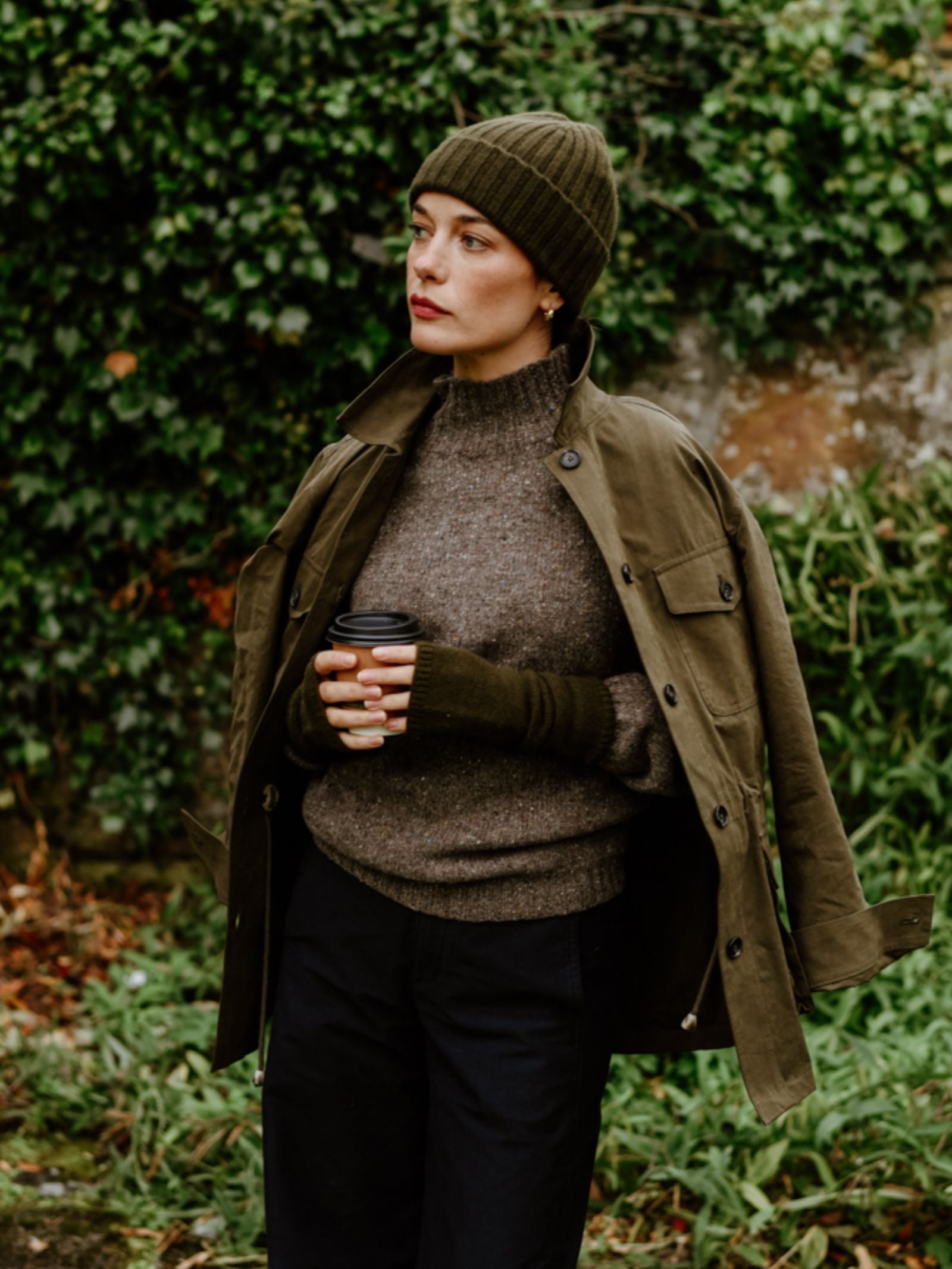 A person stands outdoors by green foliage and a stone wall, holding a coffee cup and wearing Campbell's of Beauly Cashmere Mittens, a green beanie, an olive jacket over their shoulders, a brown turtleneck sweater, and dark pants.