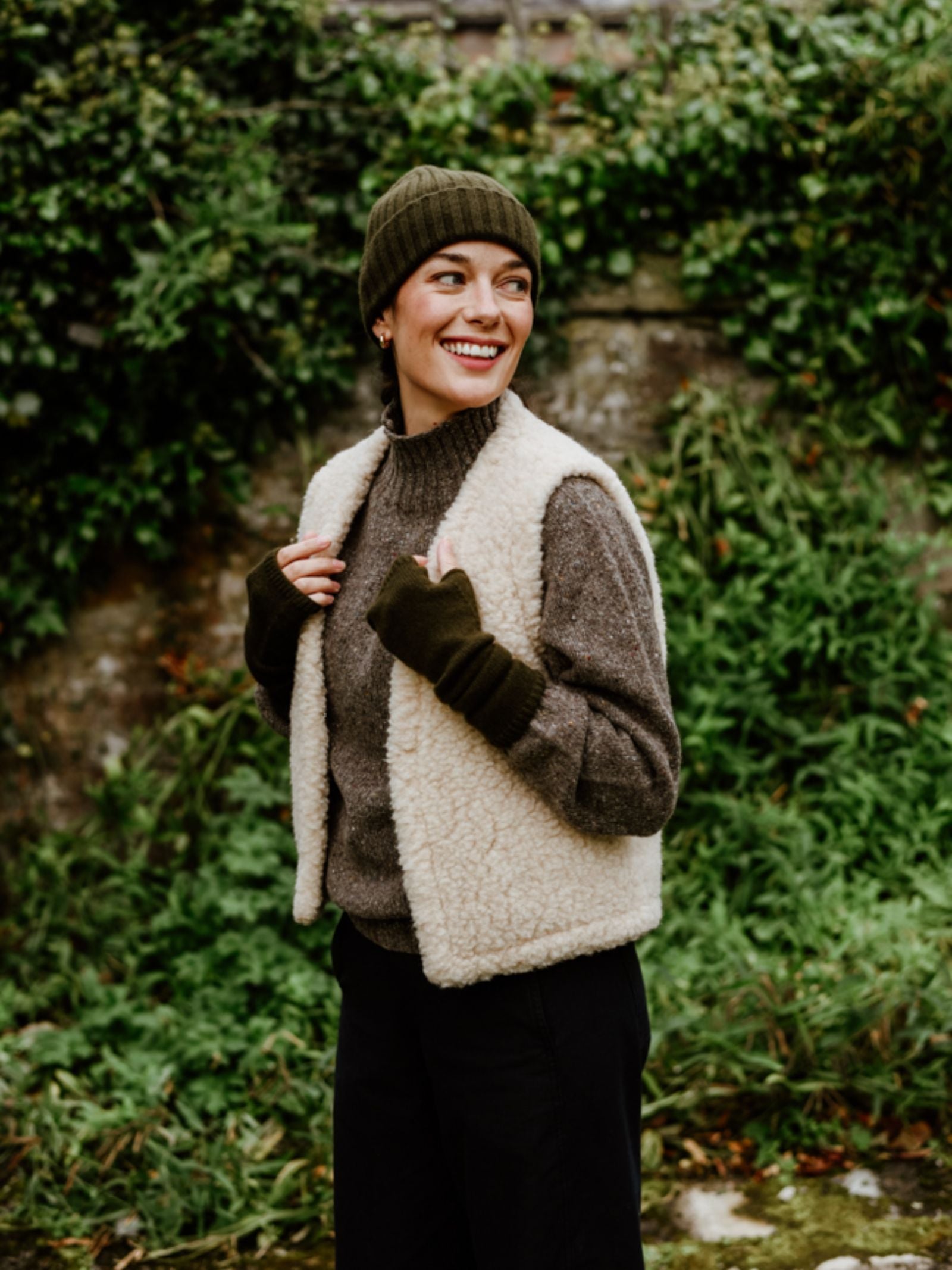 A person wearing the Campbell's of Beauly Wool Shrug smiles outdoors in front of lush green foliage, showing off lightweight warmth.