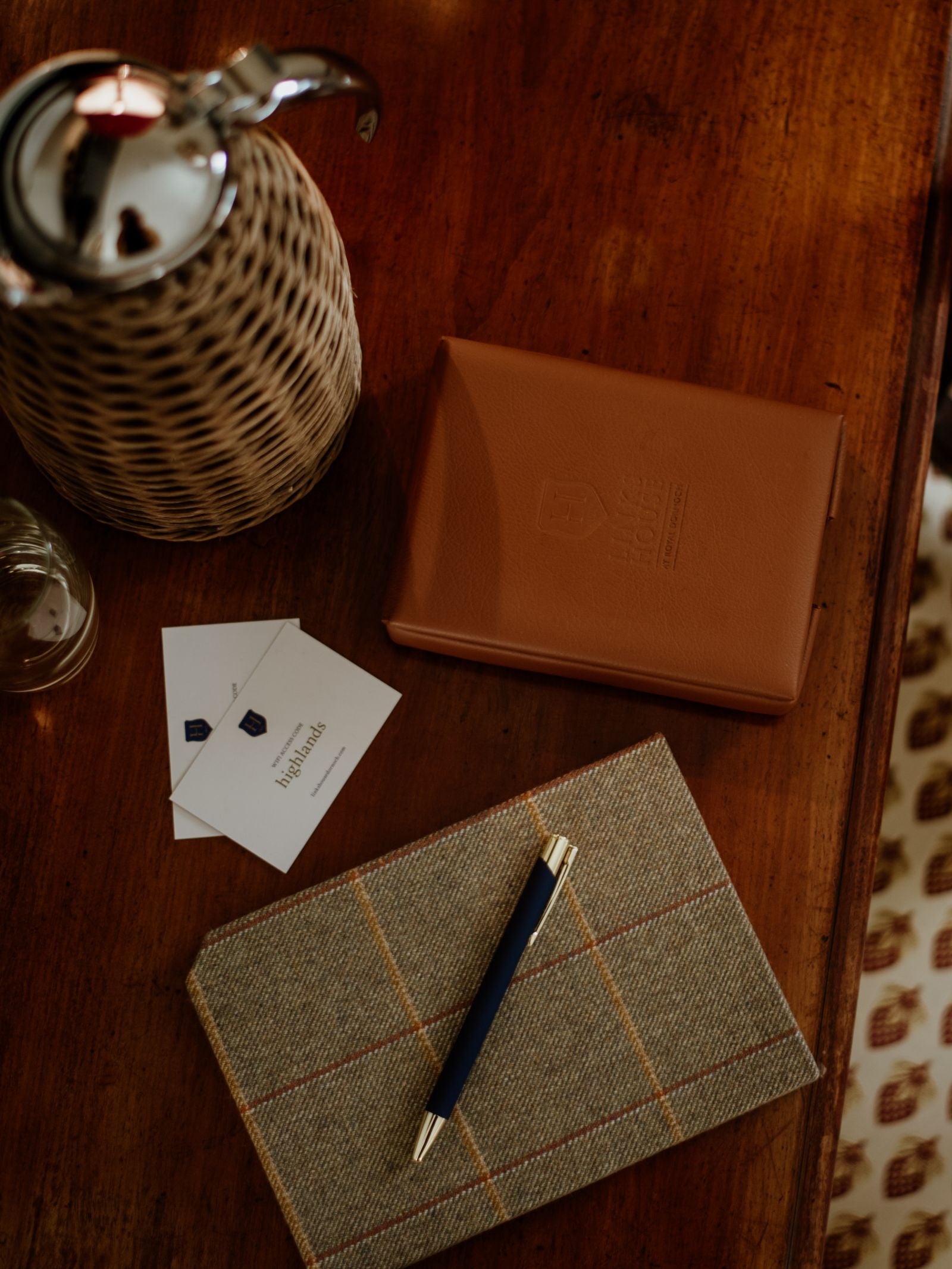 A pen rests on a Campbell's of Beauly Tweed Notebook (A5, plain paper, eco-friendly) beside business cards, a brown leather folio, and a wicker-covered carafe on a wooden table.