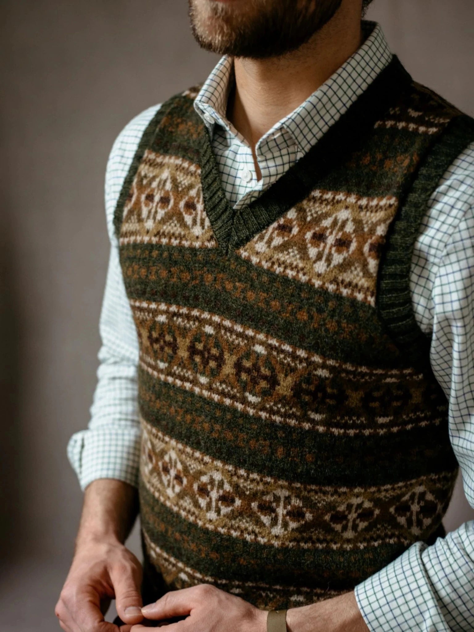 A person stands with hands loosely clasped, wearing a white checkered shirt layered with Campbell’s of Beauly’s Fairisle Slipover in earth-toned Shetland wool. The sleeveless knit adds texture, while the face is partially out of frame.