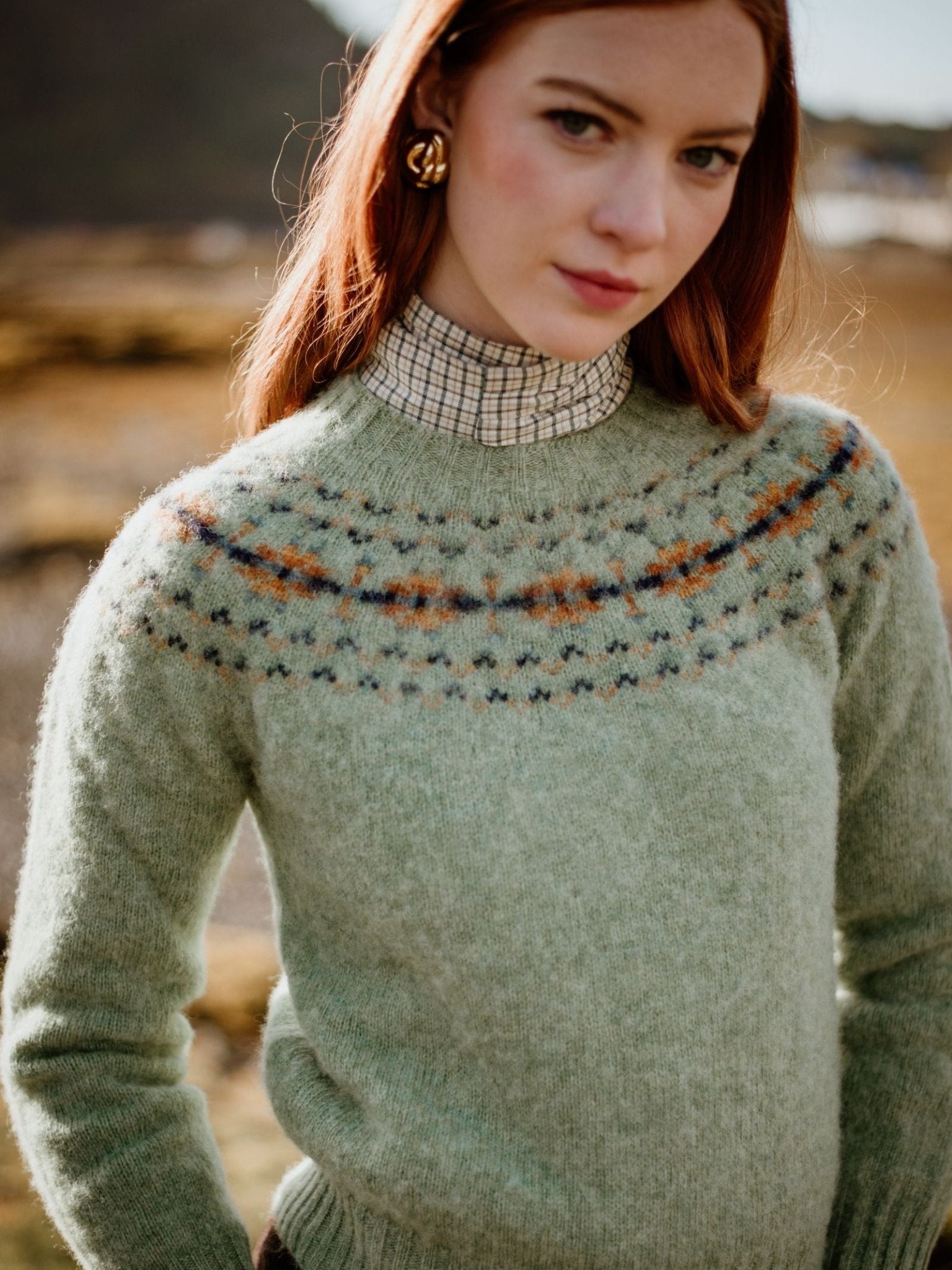 A woman with long red hair stands outdoors, looking softly to the side. She wears Campbell's of Beauly's Auld Stock Cropped Fairisle Crew Neck Jumper—green patterned knit—layered over a plaid turtleneck.