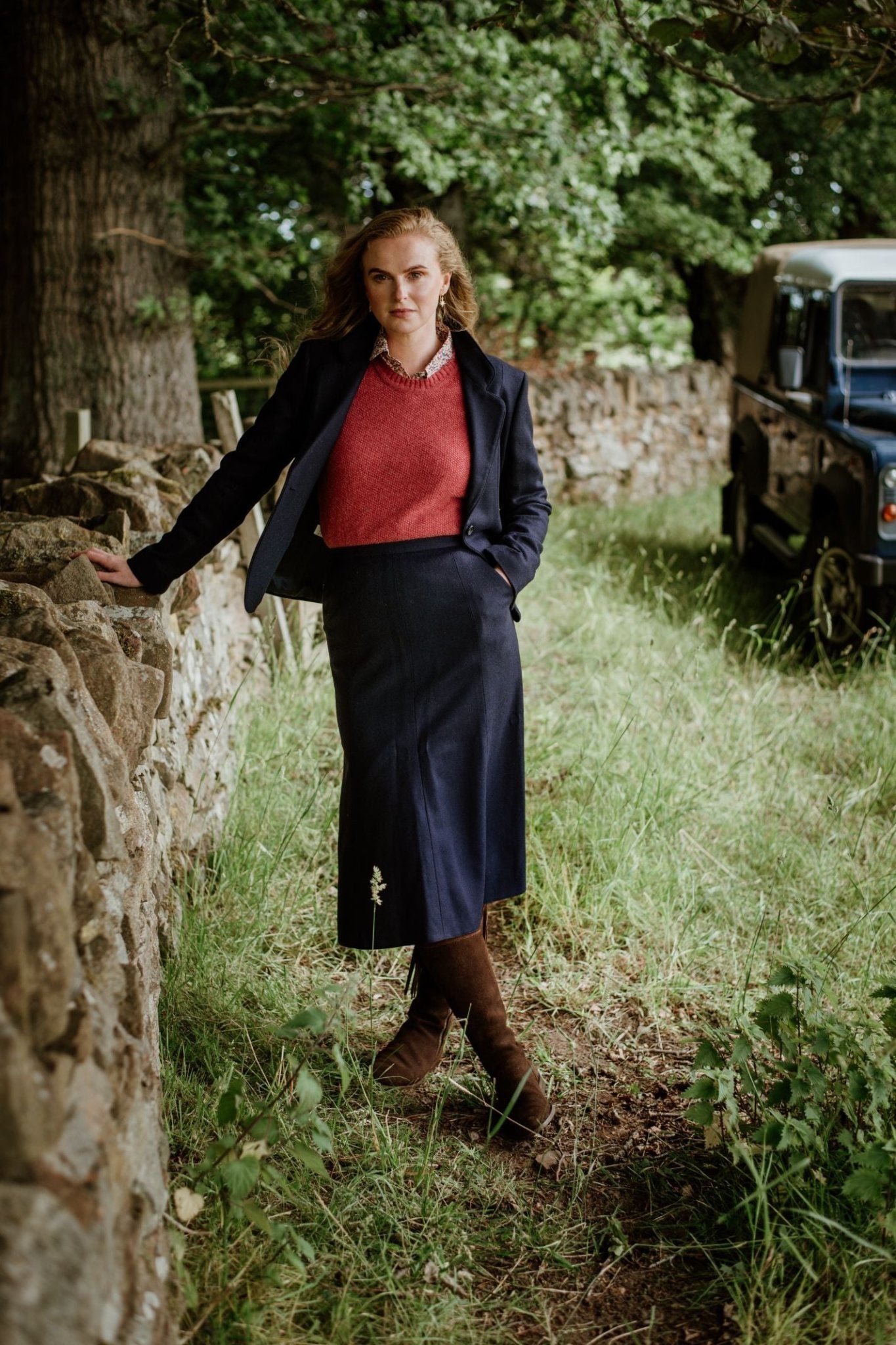 A woman stands by a stone wall outdoors, wearing Campbell's of Beauly's Panel Skirt with a navy jacket and red sweater. Brown boots complete the look. She is surrounded by grass and trees, with a blue car partly visible behind her.