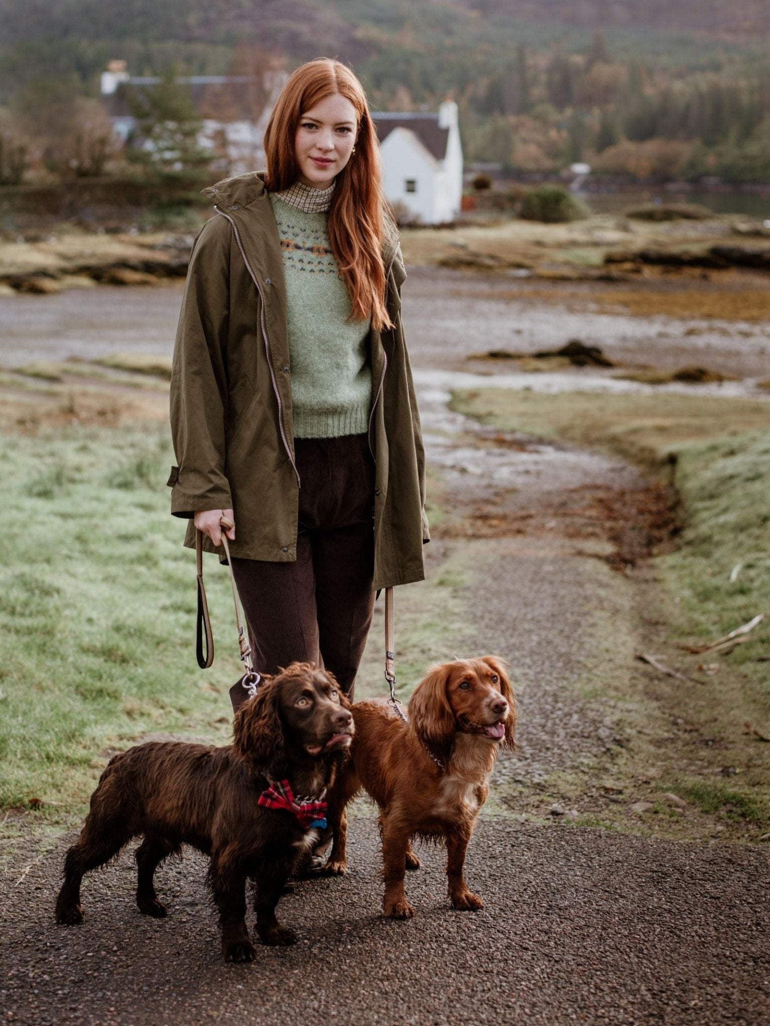 A woman with long red hair, wearing a green jacket and the Campbell's of Beauly Auld Stock Cropped Fairisle Crew Neck Jumper, stands on an outdoor path holding leashes for two brown spaniels. A white house and trees appear in the background.