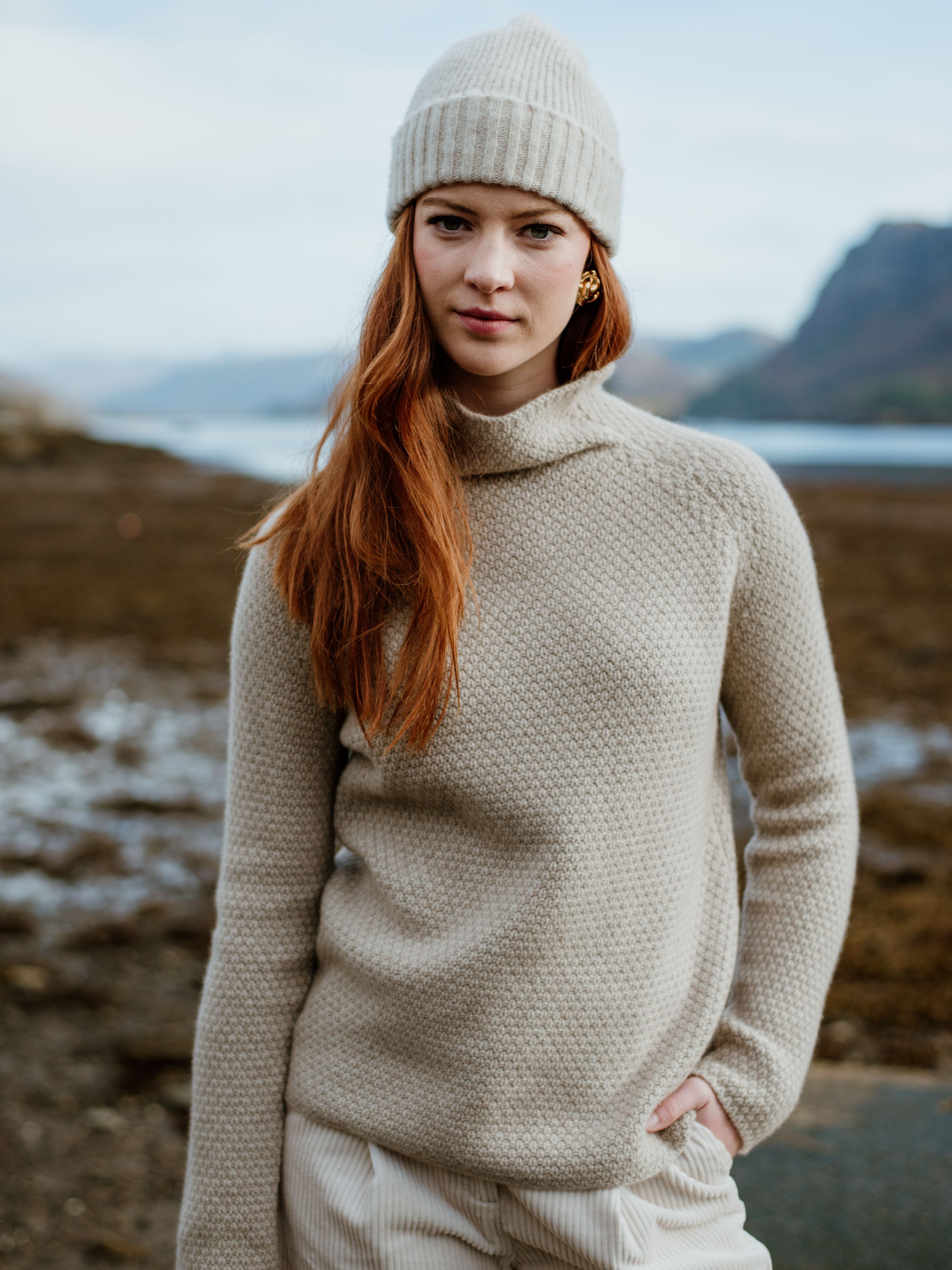 A woman with long red hair wears a cream knit beanie and sweater, standing outdoors near water and mountains, with her hand in her pocket. The background is slightly blurred, highlighting her calm expression.