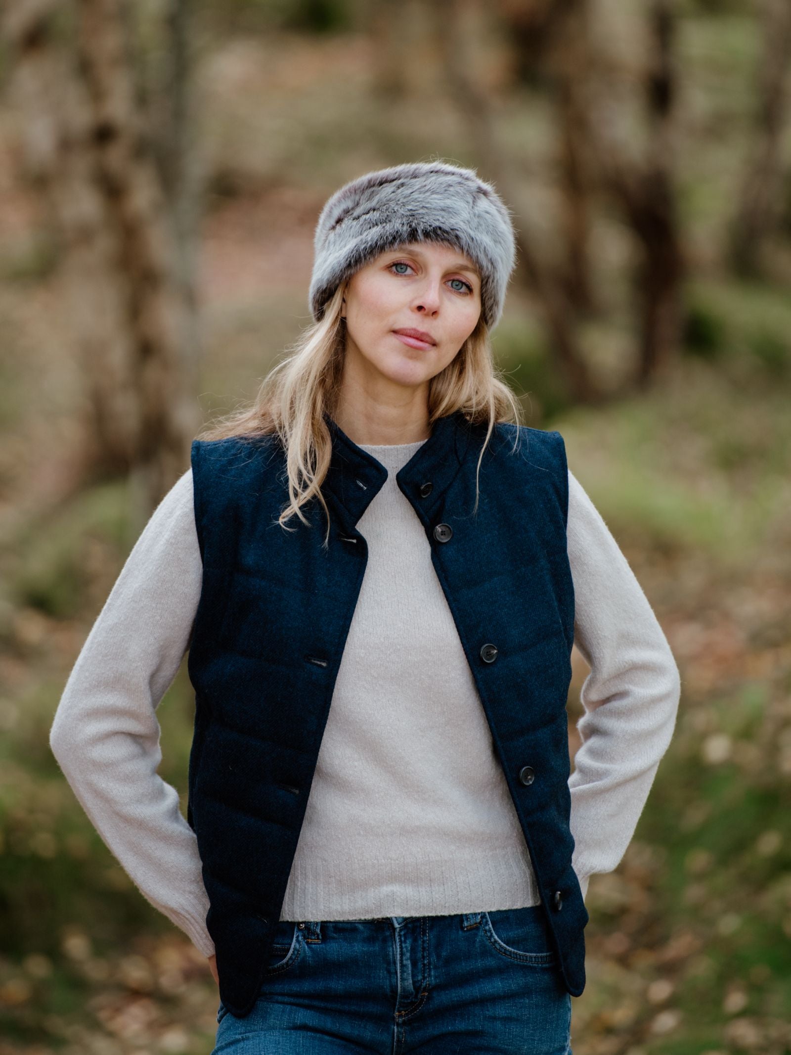 A woman with blonde hair wears the Campbell's of Beauly Sheepskin Headband, paired with a dark blue vest and cream sweater, standing outdoors.