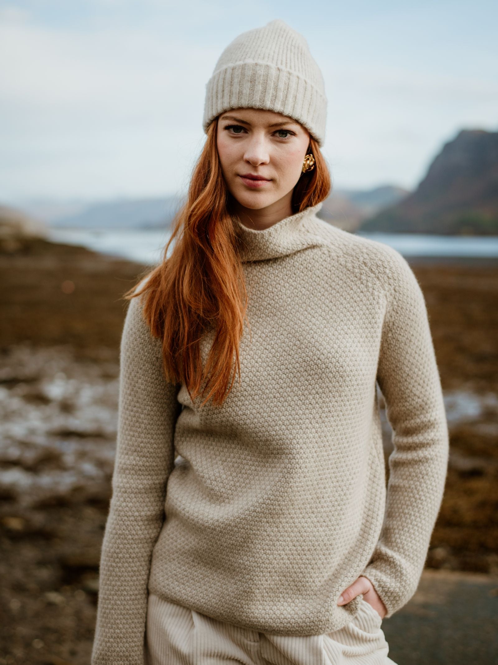 A woman with long red hair stands by the rocky Scottish Borders shoreline, calmly facing the camera. She wears a beige knit sweater and a Cream Lambswool Hat from Campbell's of Beauly, with one hand in her pocket. Mountains and water are visible behind her.