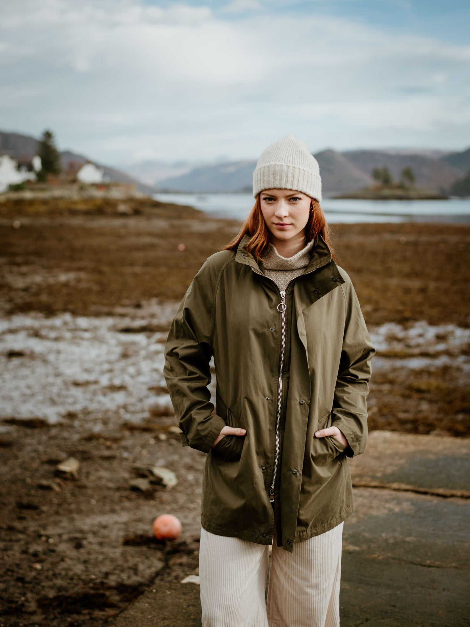 A young woman with reddish hair wears the Cream Lambswool Hat by Campbell's of Beauly, a green coat, and cream pants while standing outdoors by a rocky Scottish shore with mountains and water in the background under a partly cloudy sky.