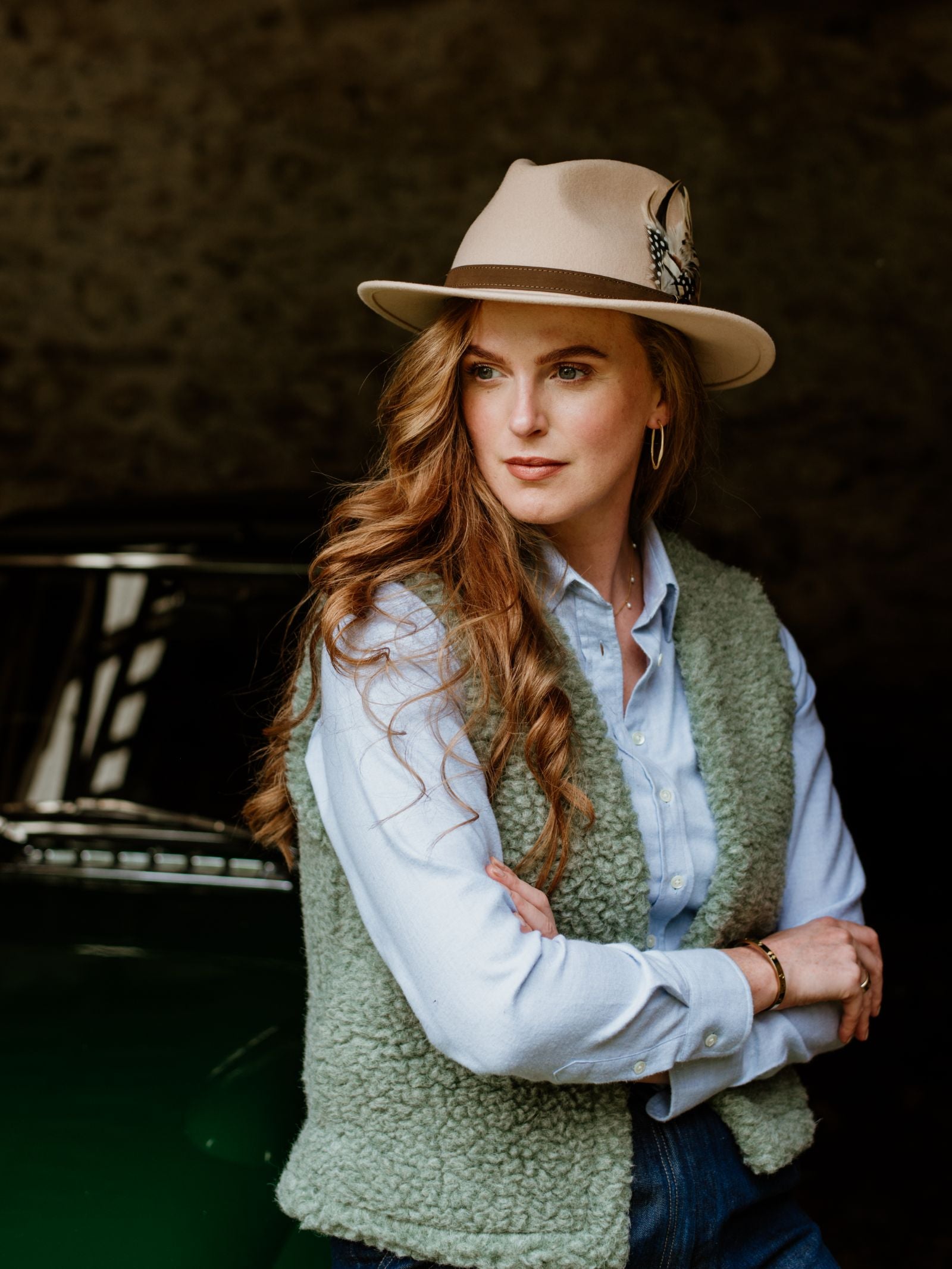 A woman with long wavy hair, dressed in a light blue shirt and green textured vest, stands arms crossed before a dark background and classic car, wearing the Hicks & Brown Auld Stock Gamebird Fedora with feather.
