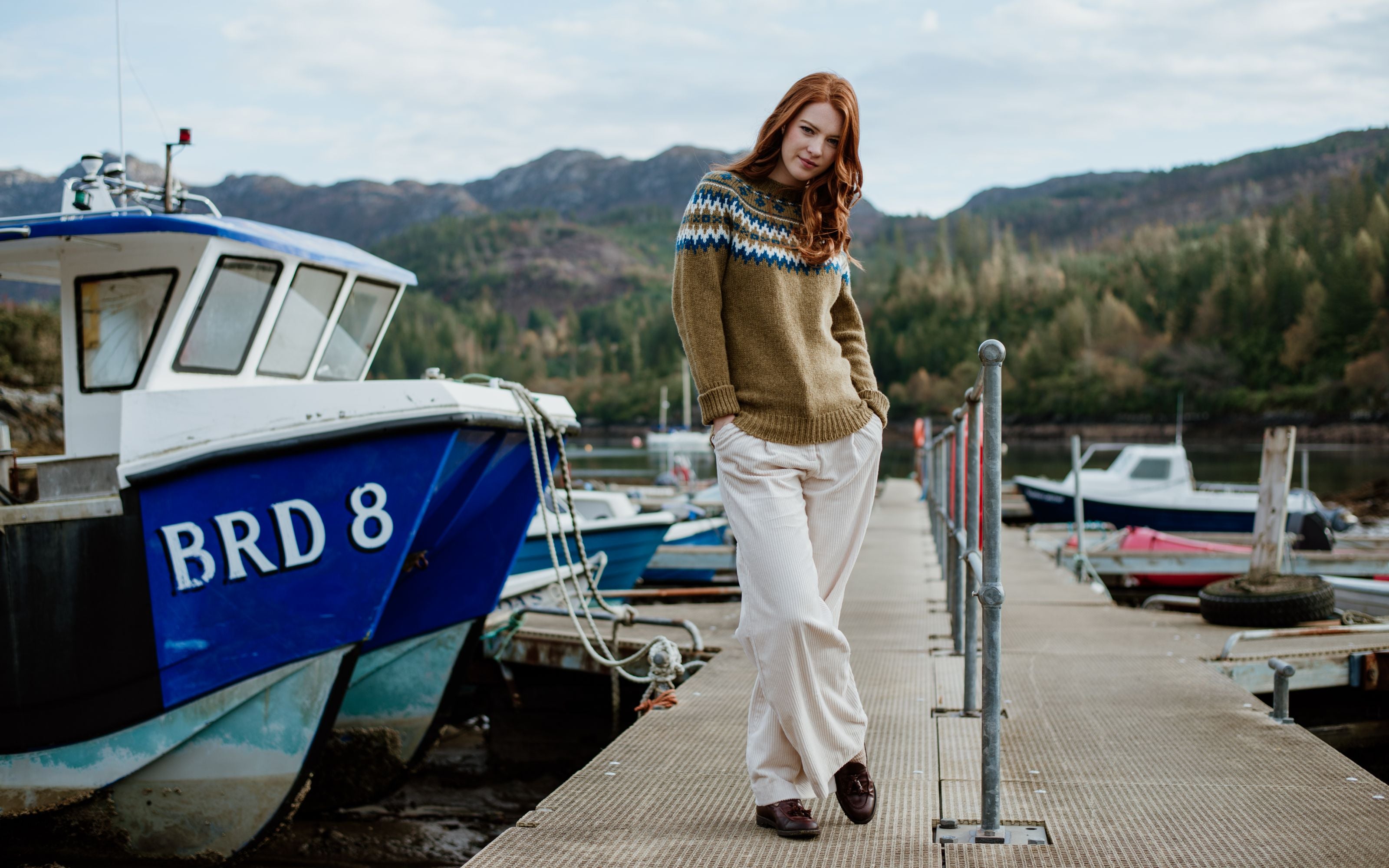 A woman with long red hair wearing a patterned sweater and light pants stands on a dock beside a blue fishing boat, with forested hills and more boats in the background.