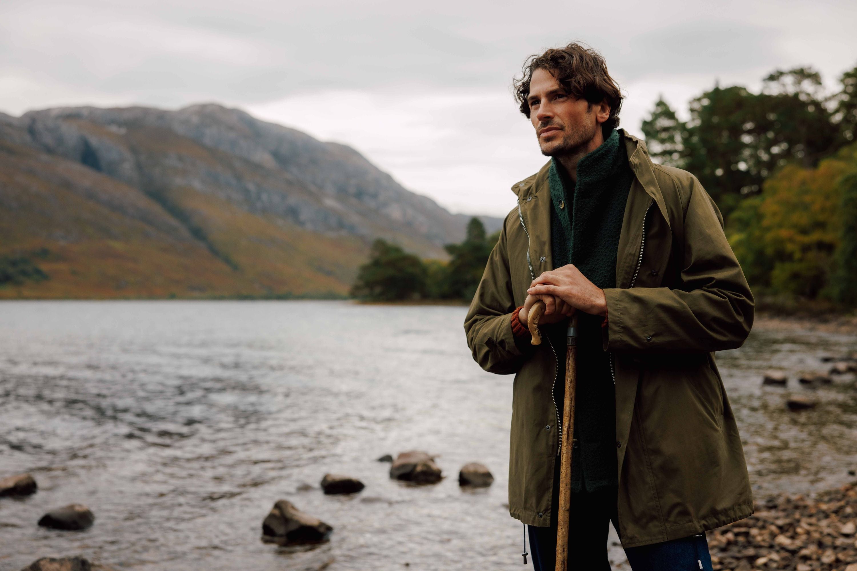 A man in an olive green jacket stands by a rocky lakeshore, holding a wooden walking stick. Mountains and trees are visible in the background under a cloudy sky.