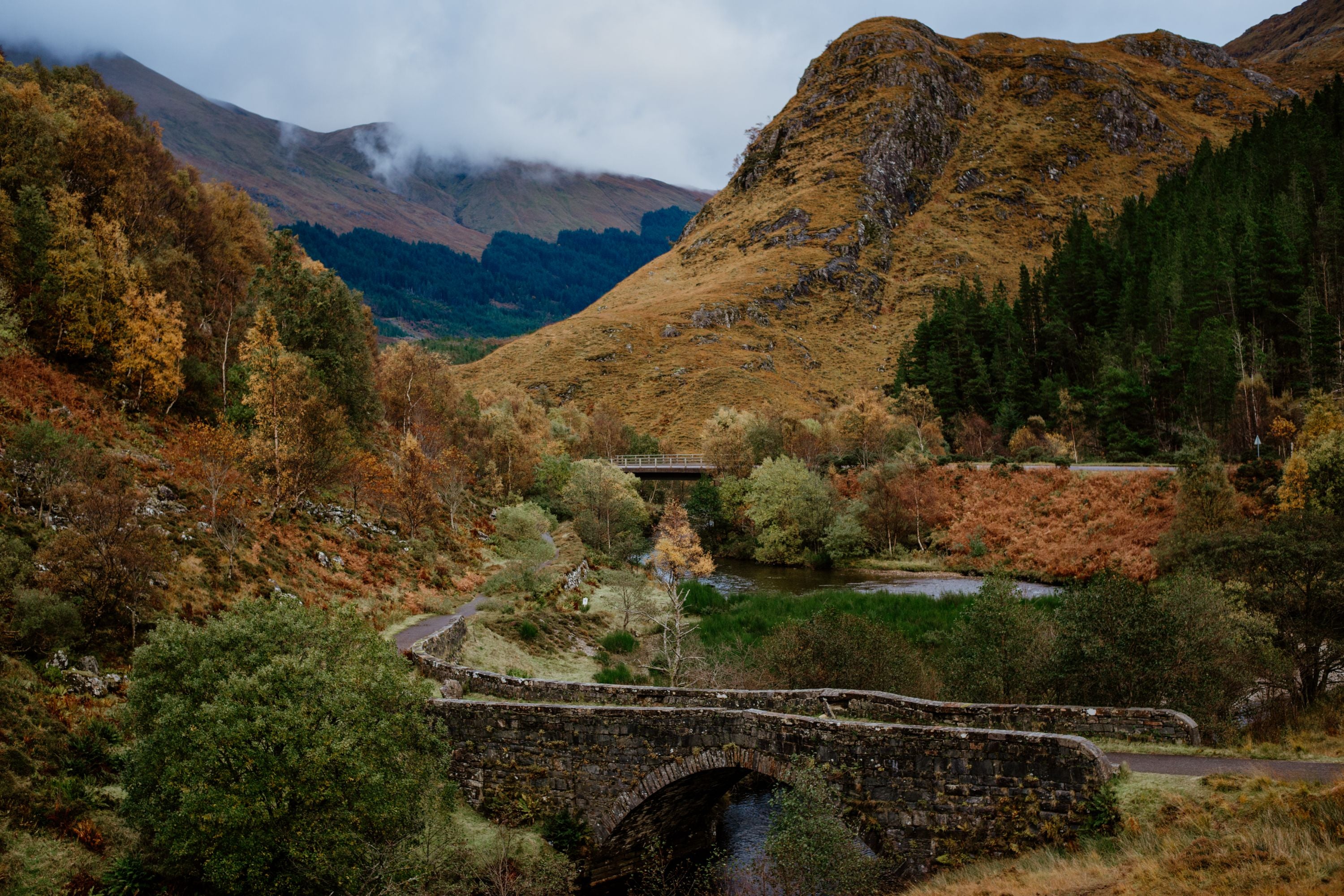 A stone bridge crosses a stream surrounded by lush trees and autumn foliage, with rolling hills and misty mountains in the background under a cloudy sky.