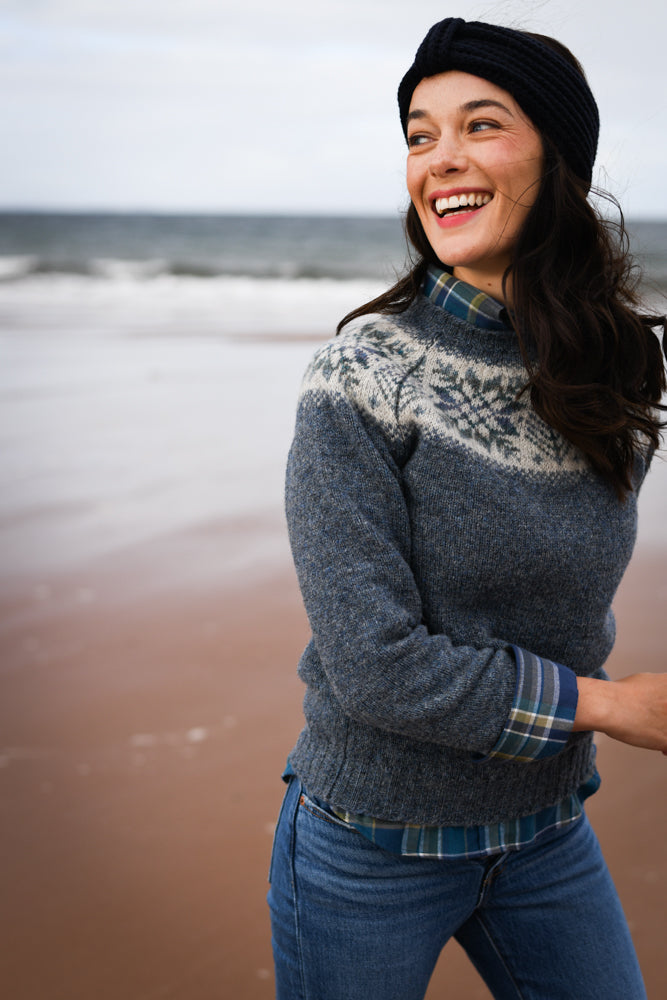 A smiling woman wearing a blue patterned sweater, plaid shirt, jeans, and a black knit headband stands on a sandy beach with the ocean and waves in the background.