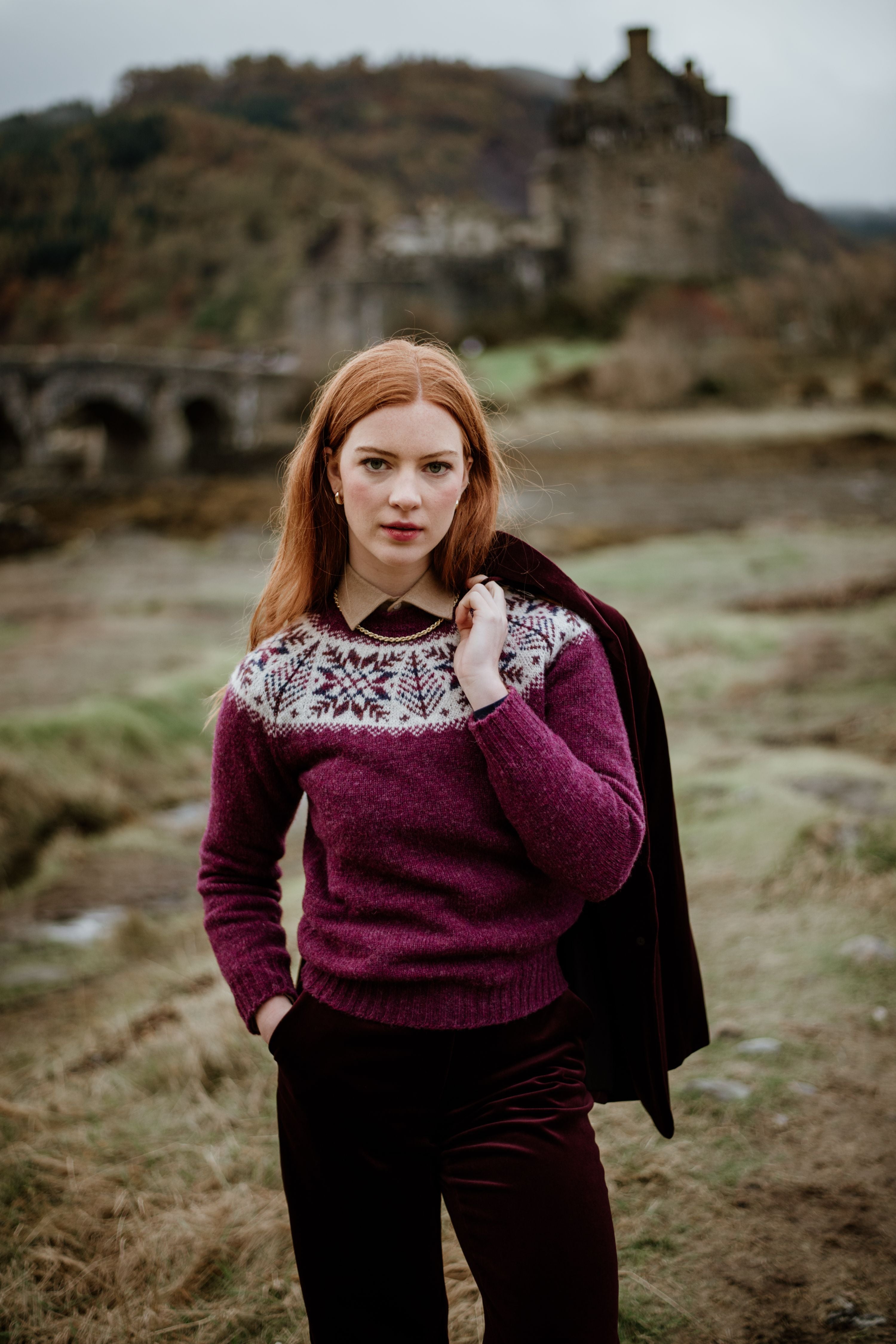 A woman with long red hair stands outdoors, wearing a purple patterned sweater and holding a dark jacket over her shoulder. A stone bridge and a castle on a hill are visible in the blurred background.