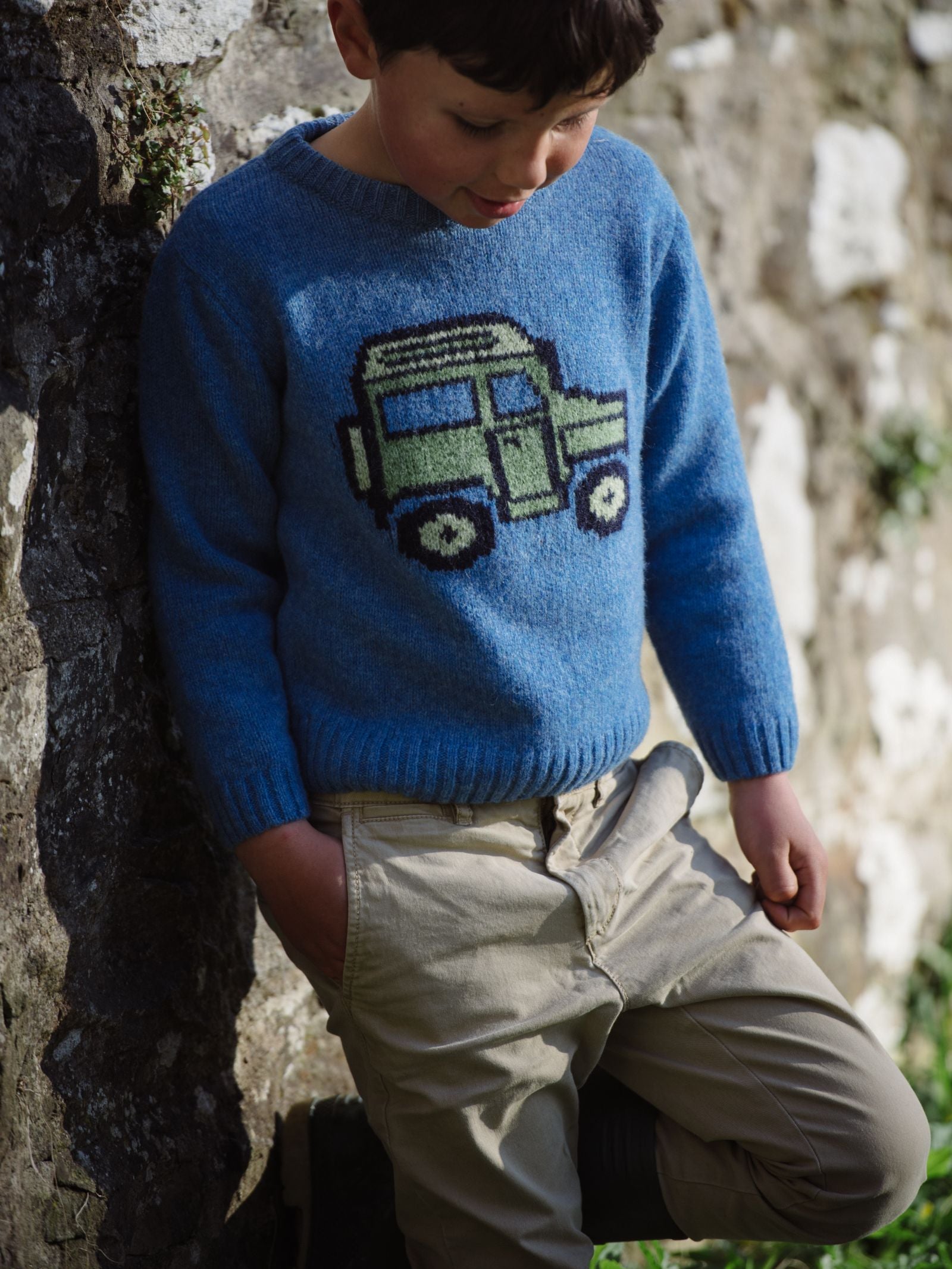 A young boy in the Campbells of Beauly Mini Land Rover Defender Jumper, featuring a green and black car design, leans against a stone wall with his hand in his pocket—a stylish tribute to classic British motoring heritage.