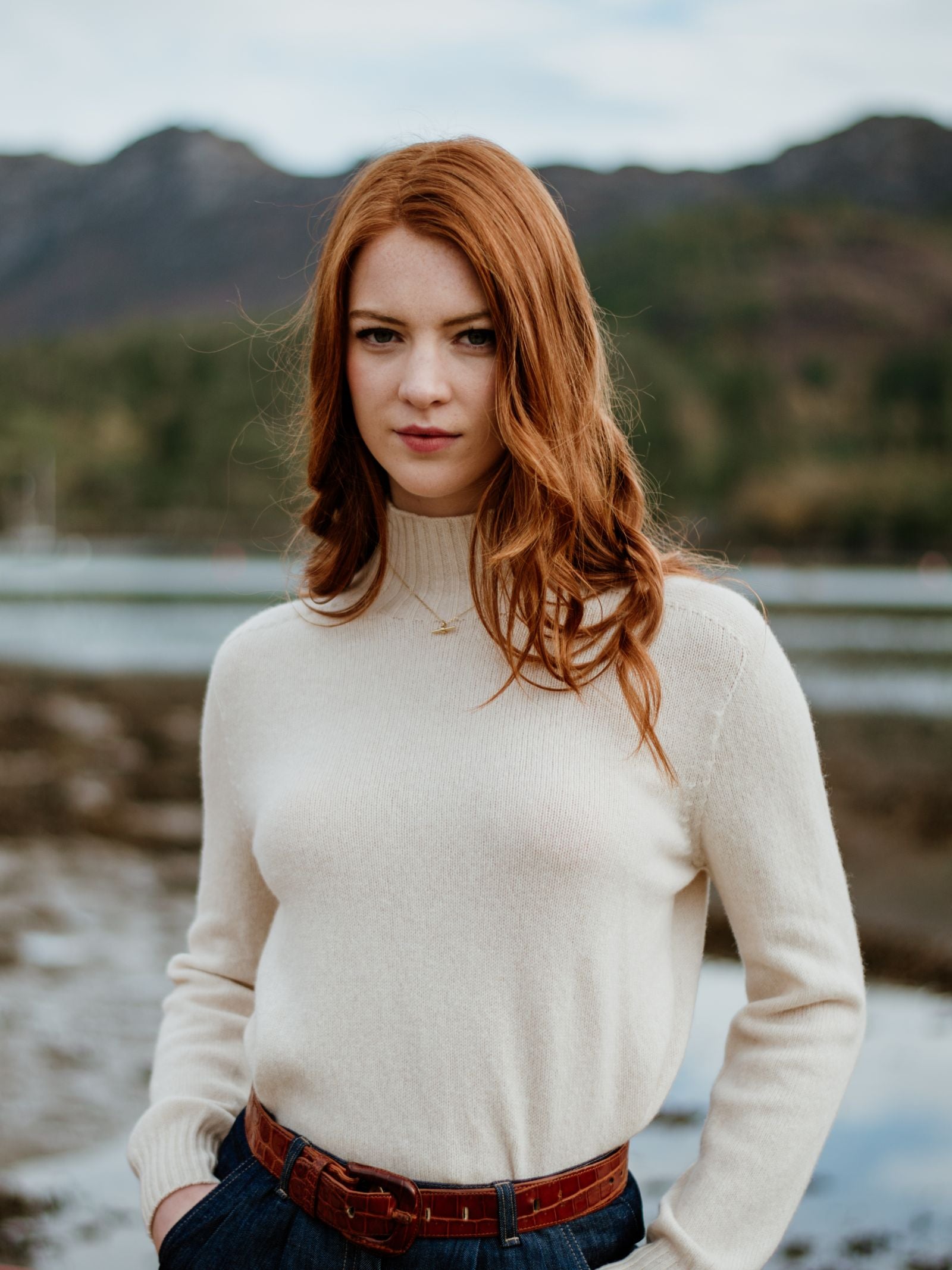 A woman with long red hair wears the Campbells of Beauly Geelong Lambswool Funnel Neck sweater outdoors by water, with blurred mountains and trees behind her. She gazes calmly at the camera.