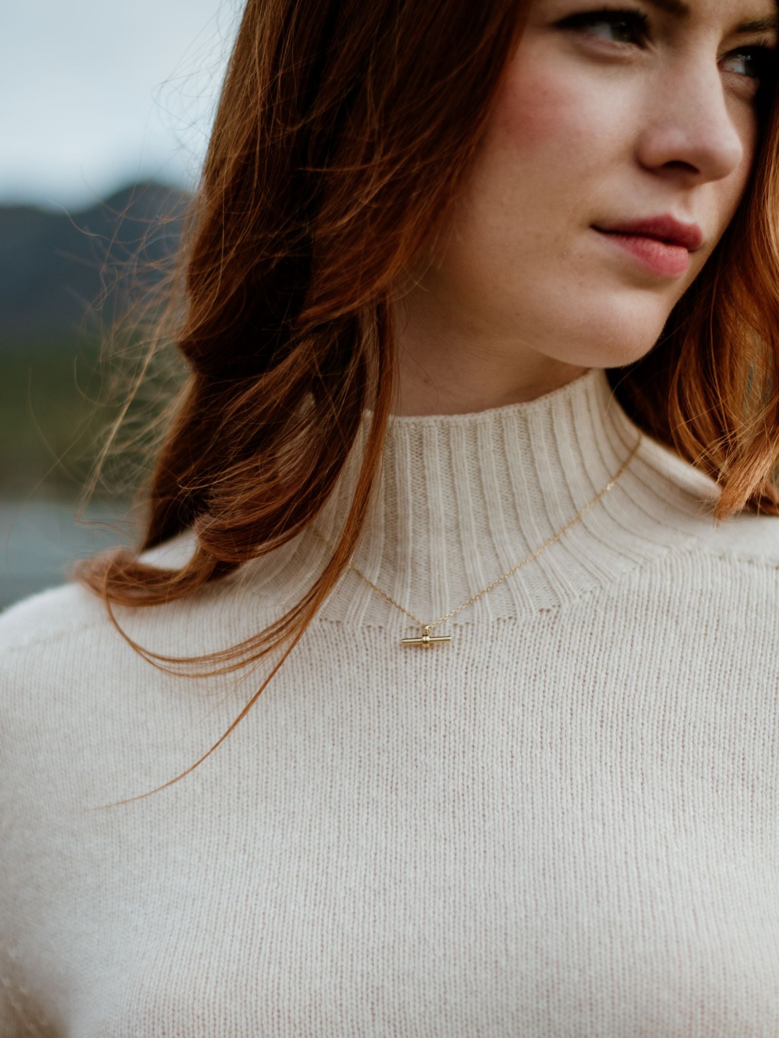 A woman with long red hair wears a cream-colored Campbell’s of Beauly Geelong Lambswool Funnel Neck sweater and a delicate gold necklace. The photo is cropped to her lower face and chest, set against a blurred outdoor background.