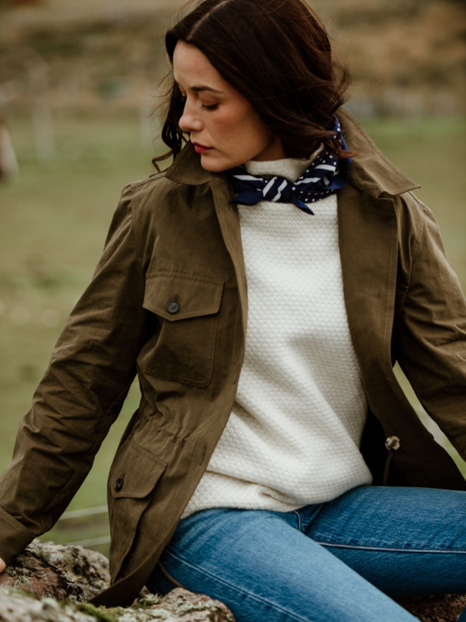 A dark-haired woman looks down, wearing the Campbell's of Beauly Auld Stock Aigas Field Jacket in water-repellent peach skin over a white sweater and blue jeans, accessorized with a blue and white patterned scarf.