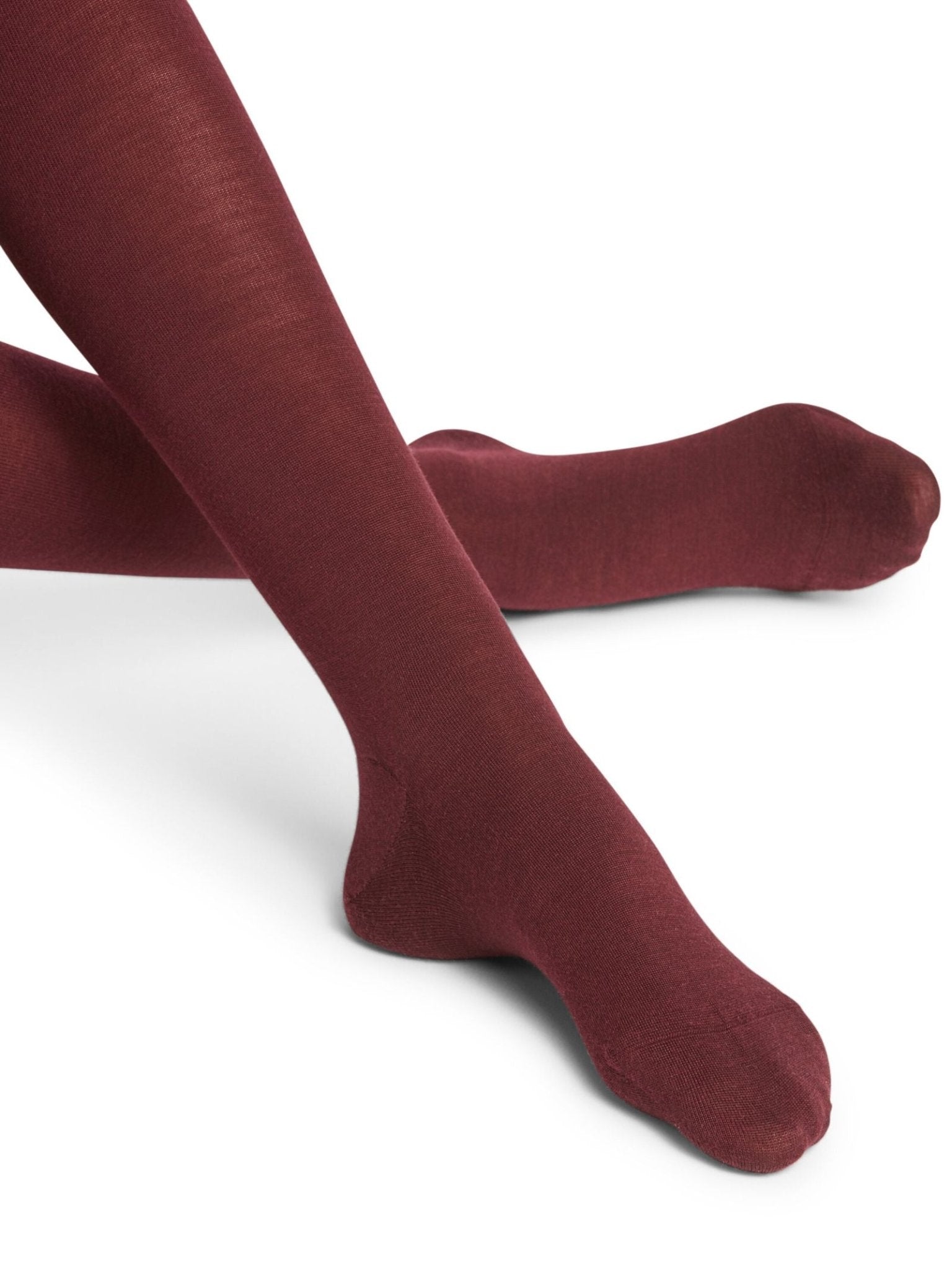 Close-up of crossed legs wearing FALKE Merino Tights in opaque burgundy, set against a plain white background.