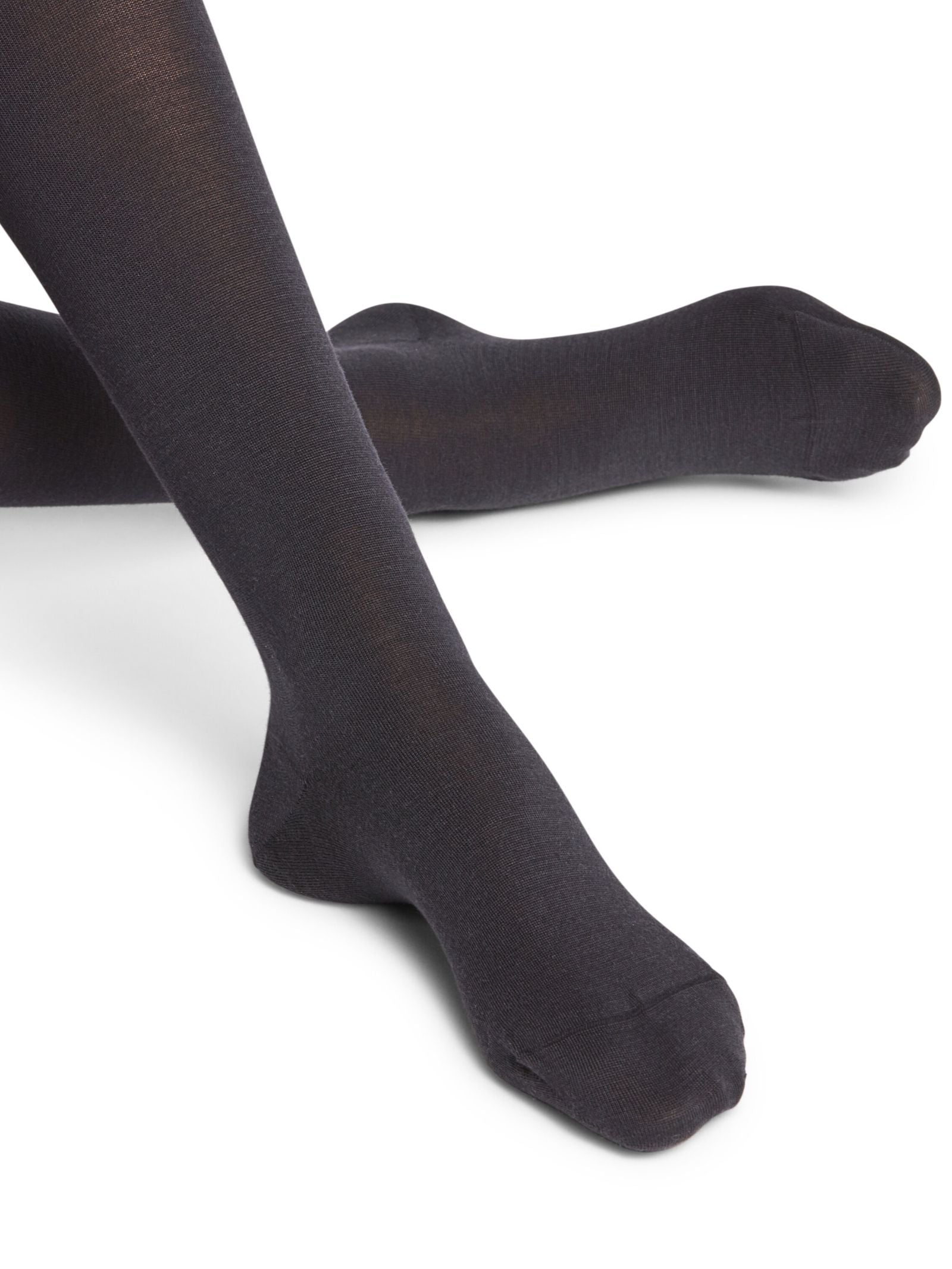 A close-up of a person’s ankles crossed, wearing FALKE Merino Tights in opaque black against a white background.