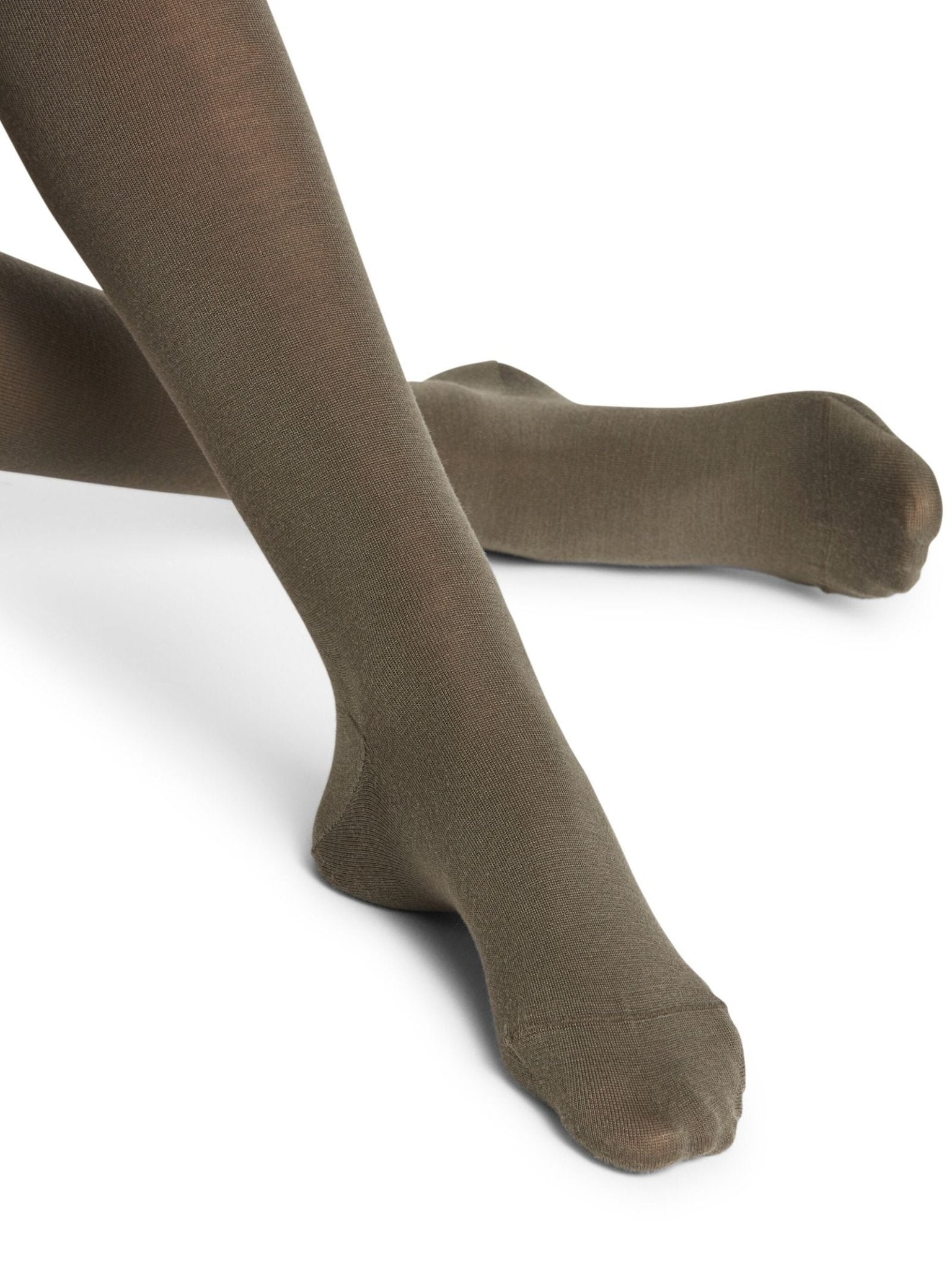 A person sits cross-legged on a white background wearing opaque dark brown FALKE Merino Tights, which are made from soft merino wool and provide full coverage to the feet and legs, with their toes slightly pointed.