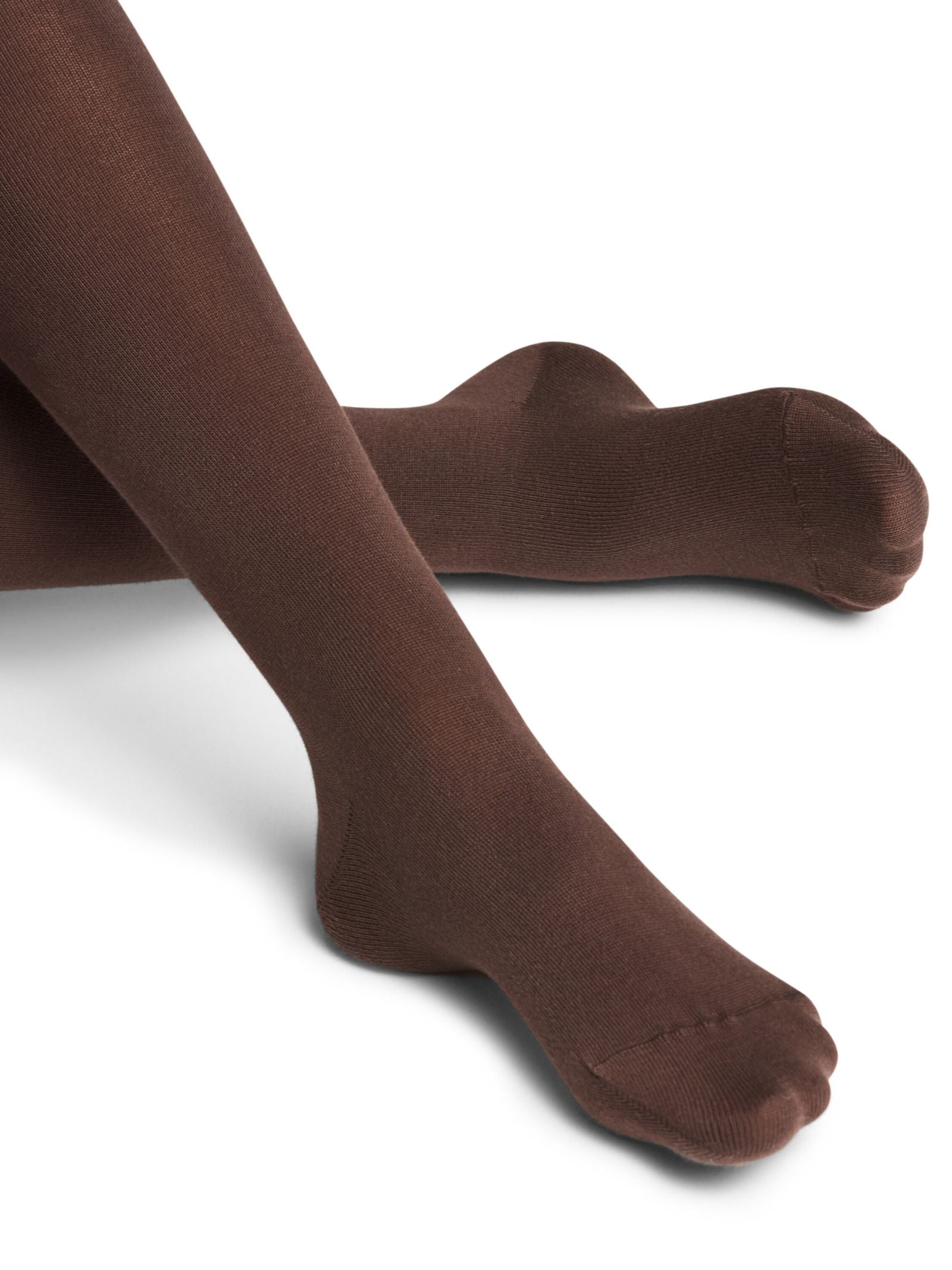 A close-up of crossed legs and pointed toes wearing FALKE Cotton Tights in opaque brown, set against a white background.