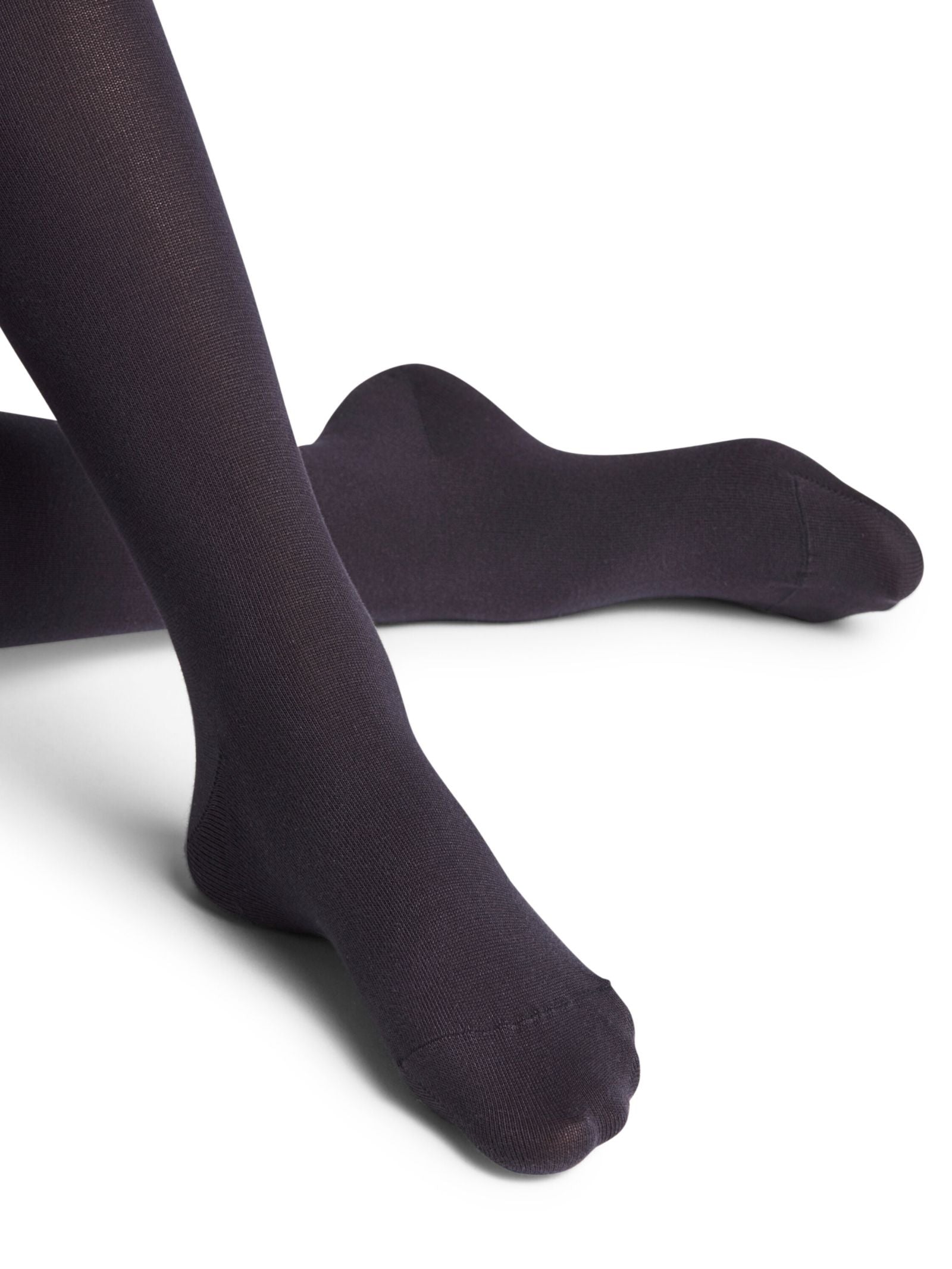 A person wears FALKE Cotton Tights, shown in opaque black with crossed legs on a white background. Only the legs and feet are visible, emphasizing the comfort and style of these FALKE sustainable cotton tights.