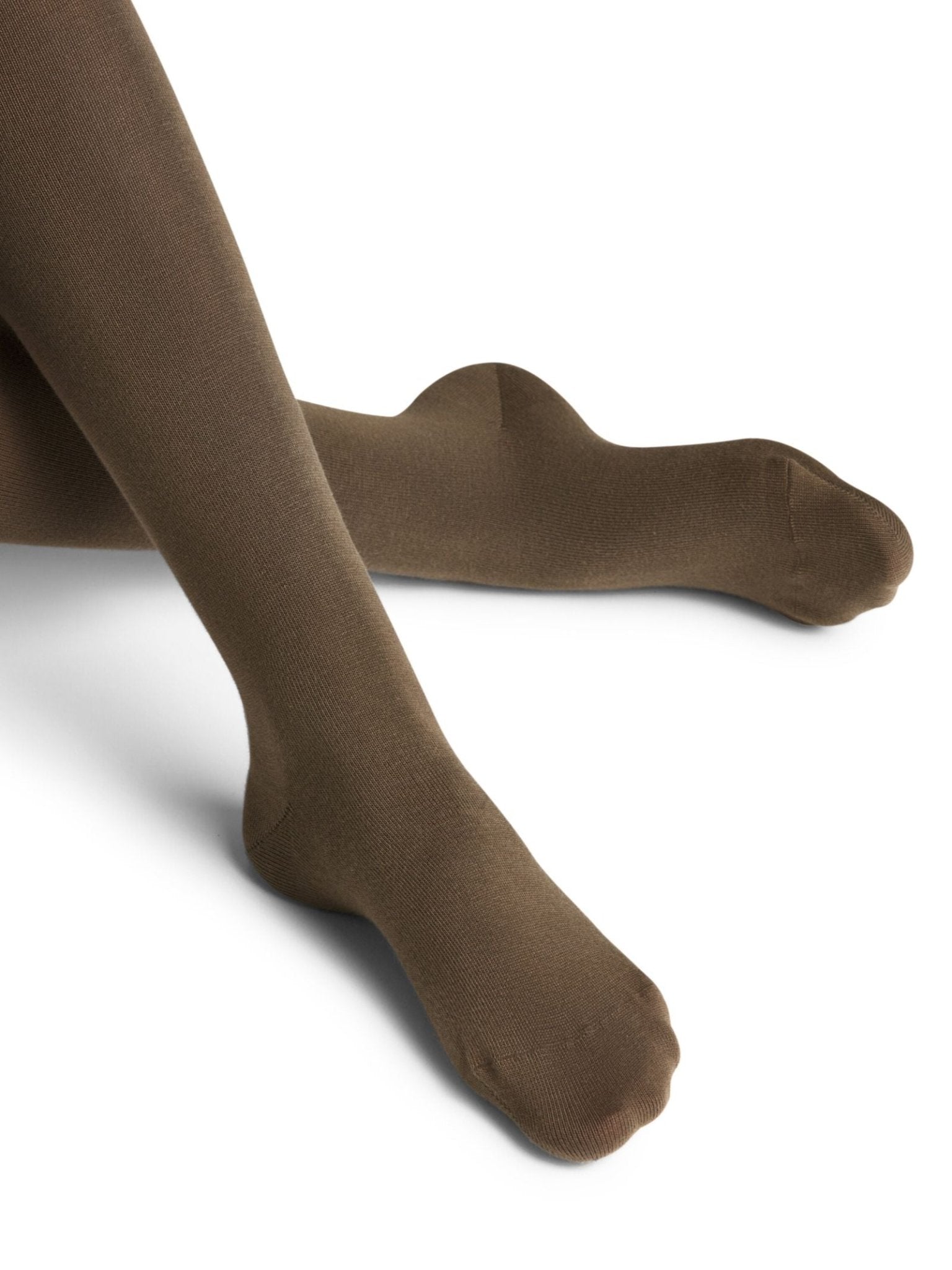 A person wearing FALKE Cotton Tights in dark brown sits cross-legged on a white background. Only their legs and feet, clad in these sustainable cotton tights, are visible.