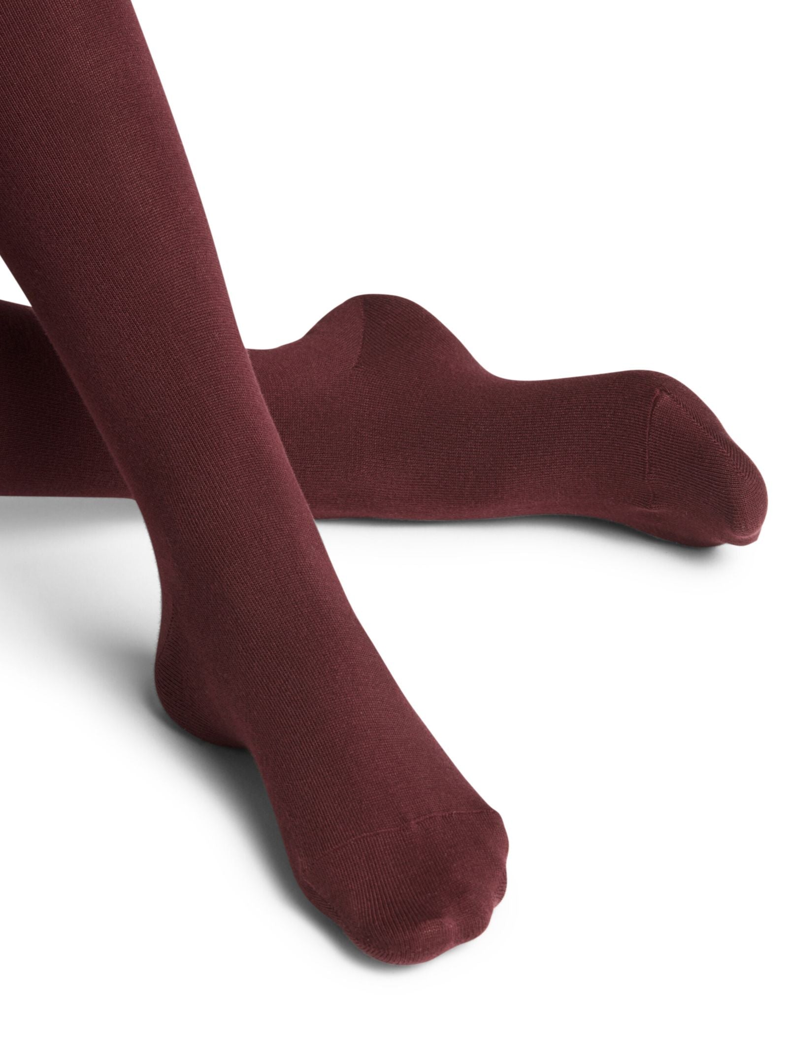 A person models FALKE Cotton Tights in opaque burgundy, sitting with one leg extended and the other bent against a white background.
