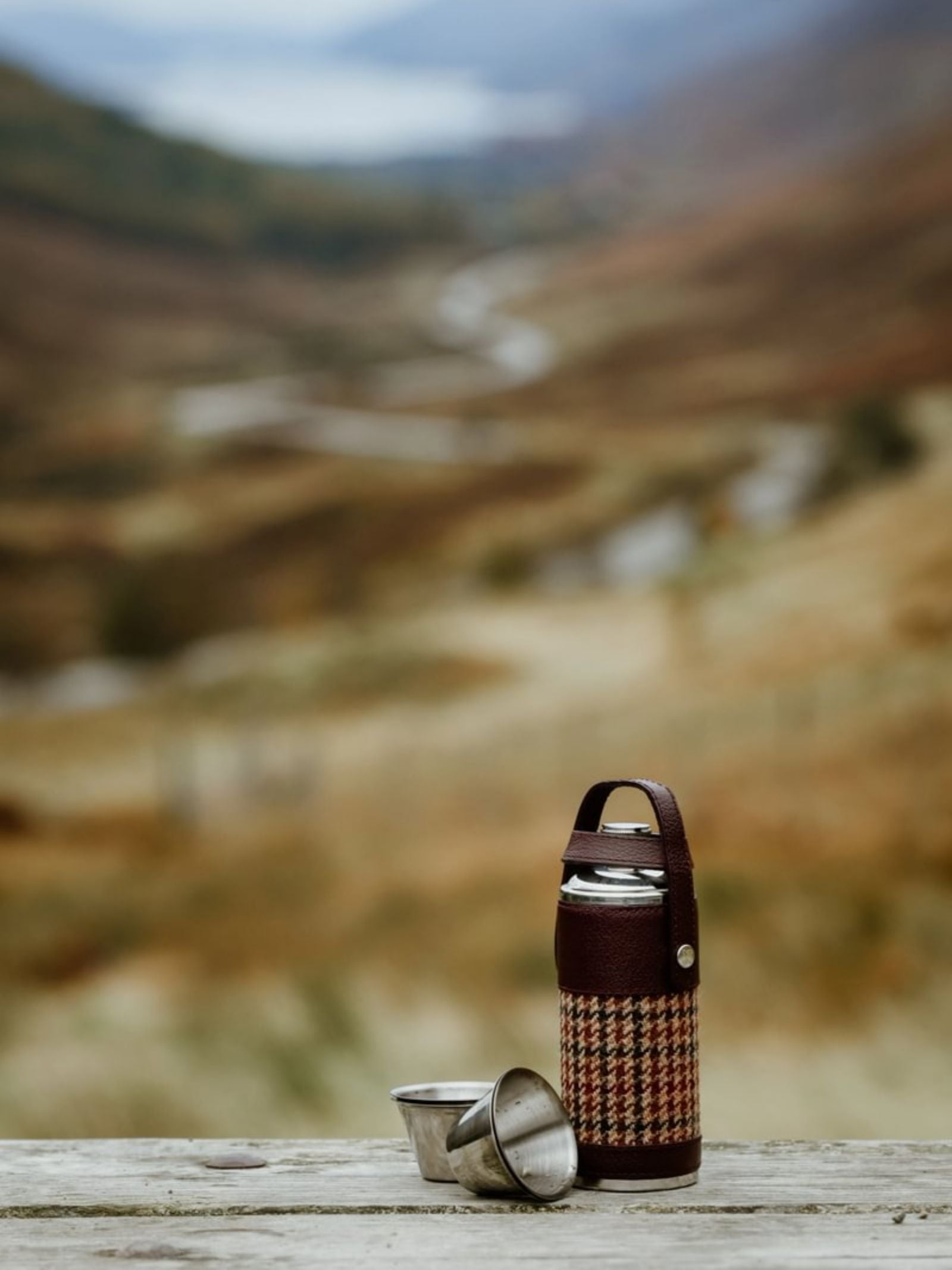 The 8oz Bordeaux Hunter Flask by Campbell's of Beauly x Ettinger, wrapped in houndstooth Capra goat leather and paired with two cups, rests on wood outdoors with rolling hills and a winding road evoking British craftsmanship.
