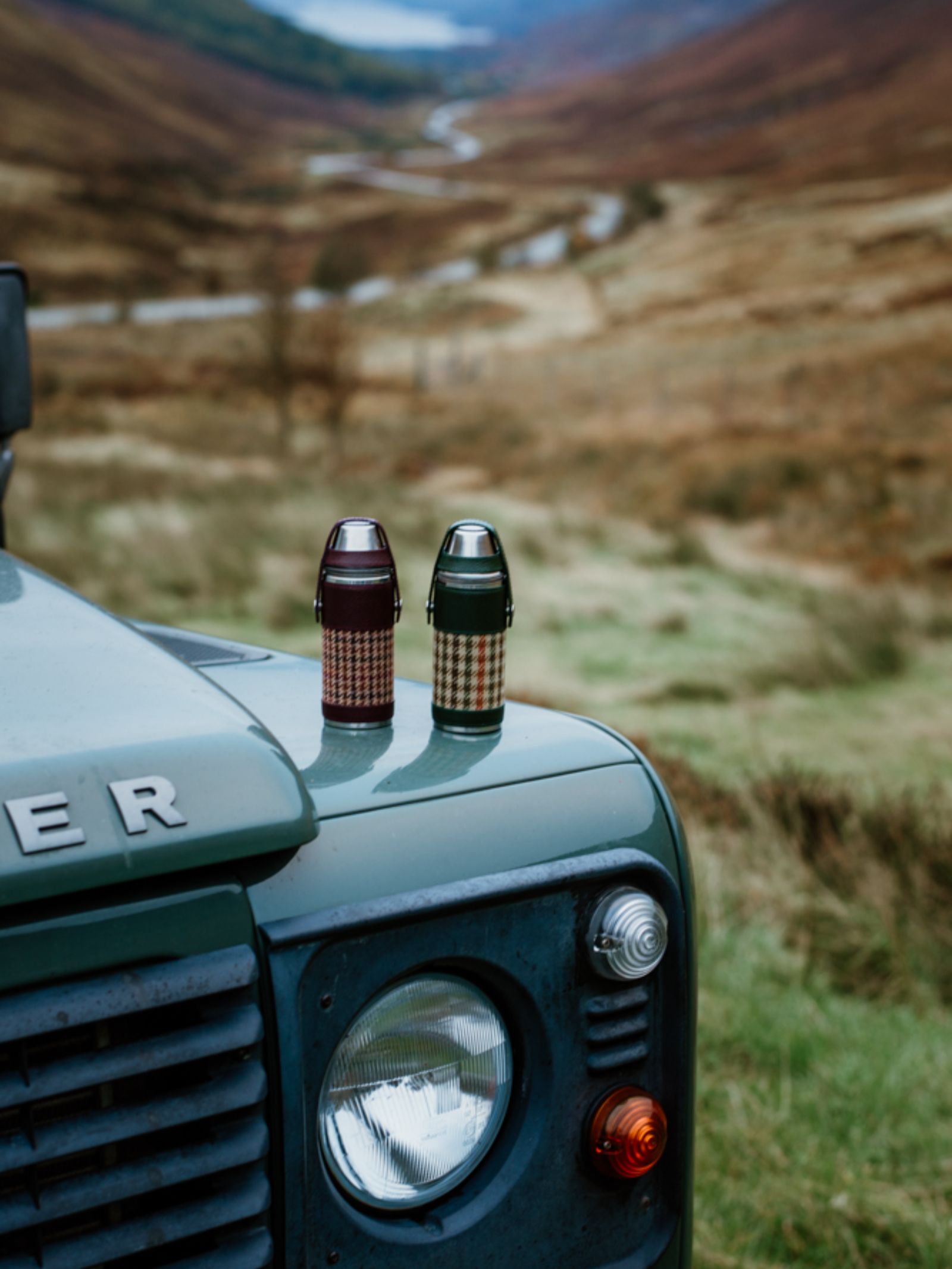 Two Campbell’s of Beauly x Ettinger 8oz Forest Green Hunter Flasks sit on a green off-road vehicle’s hood, set against hills and a winding river—highlighting British craftsmanship amidst natural beauty.