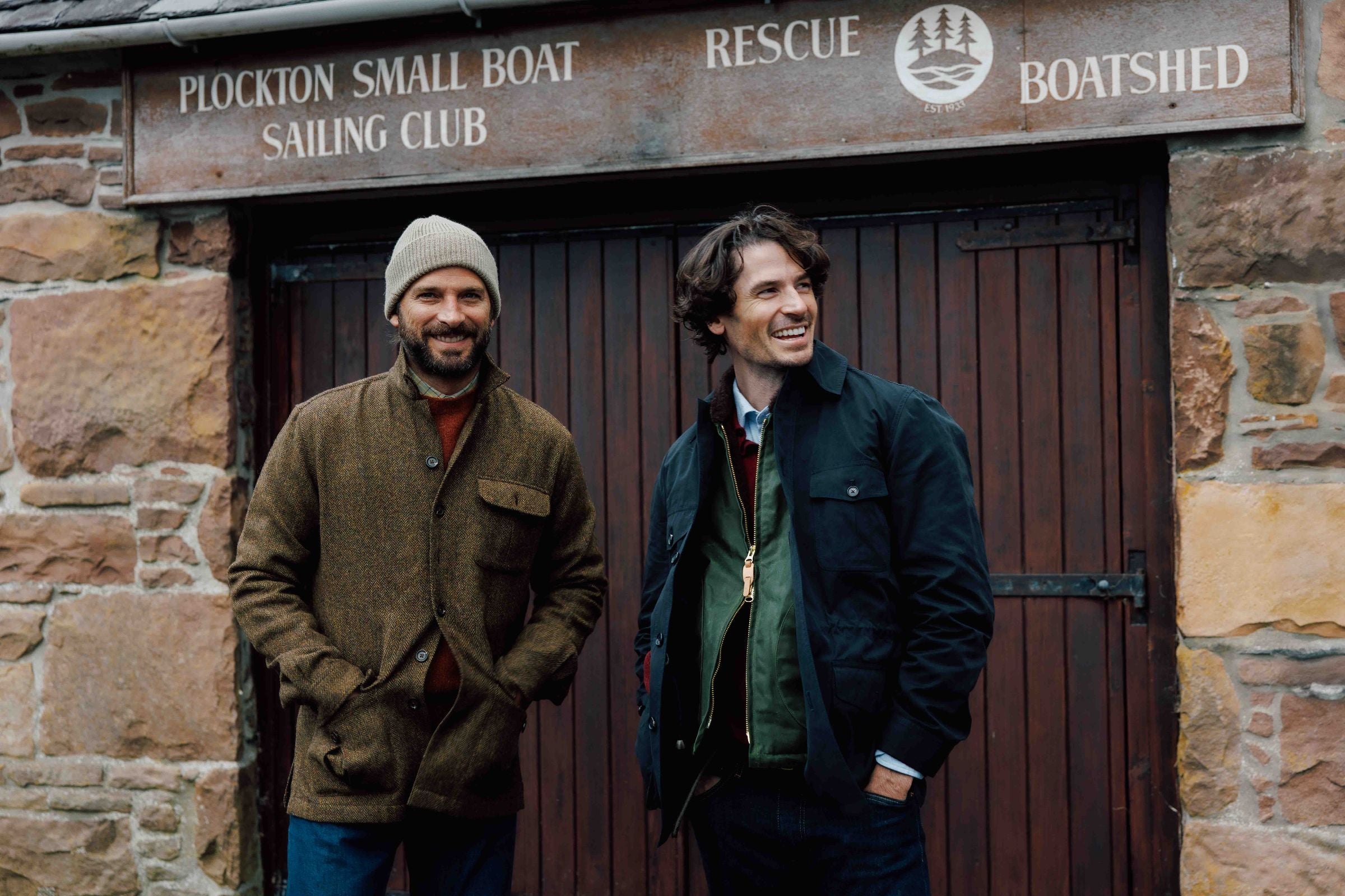 Two men in jackets stand and smile in front of a wooden door under a sign that reads Plockton Small Boat Sailing Club Rescue Boatshed. The background is made of stone bricks.
