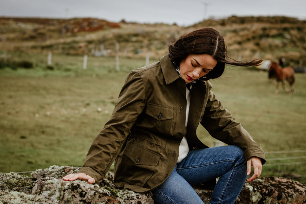 A woman in a green jacket and blue jeans sits on a rock in a grassy rural field, looking down with her hair blowing in the wind. A horse grazes in the blurred background.