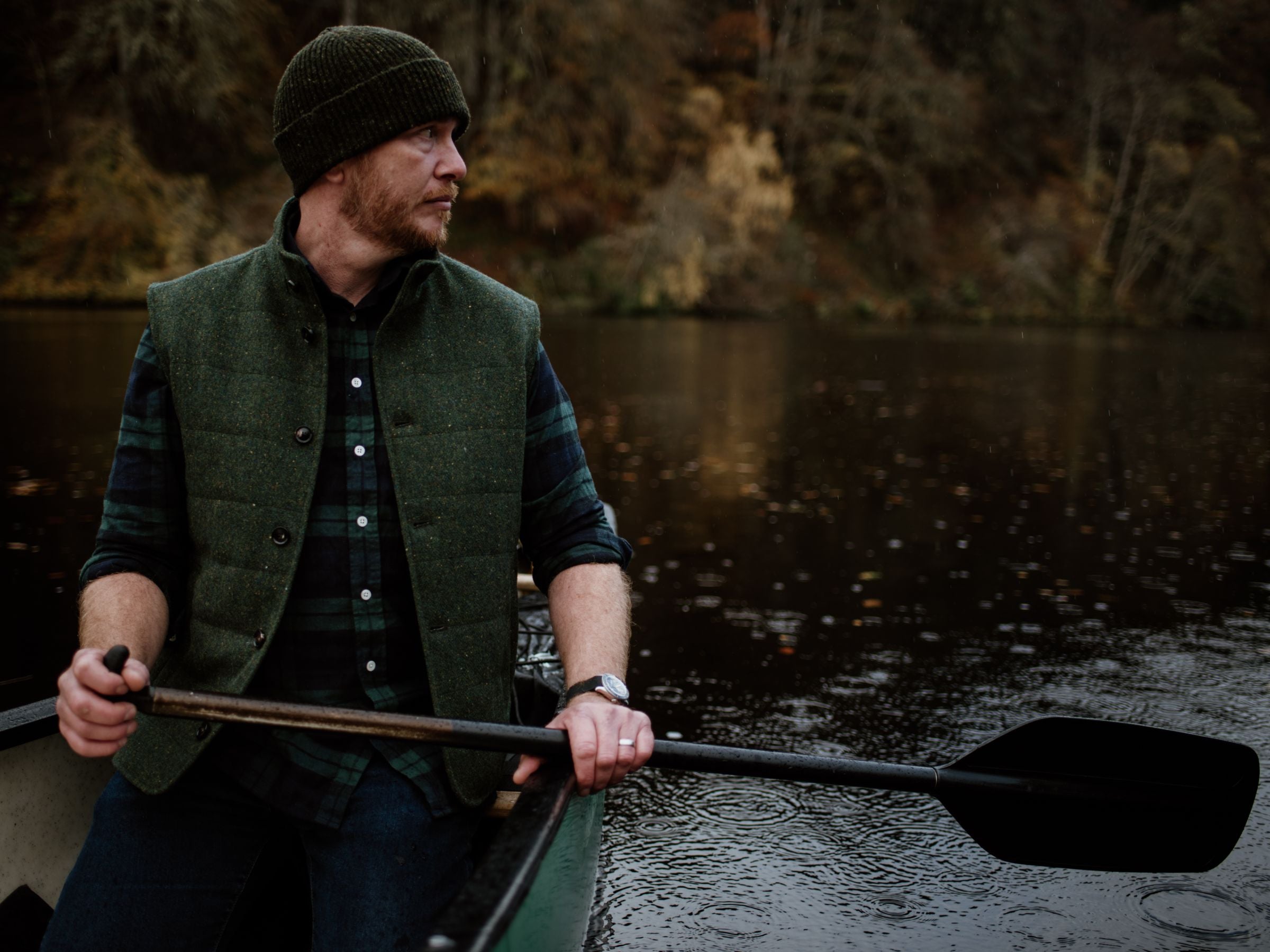 A man wearing a green knit hat and vest rows a canoe on a calm, dark lake surrounded by trees with autumn foliage. He looks to his left, holding a paddle with both hands.