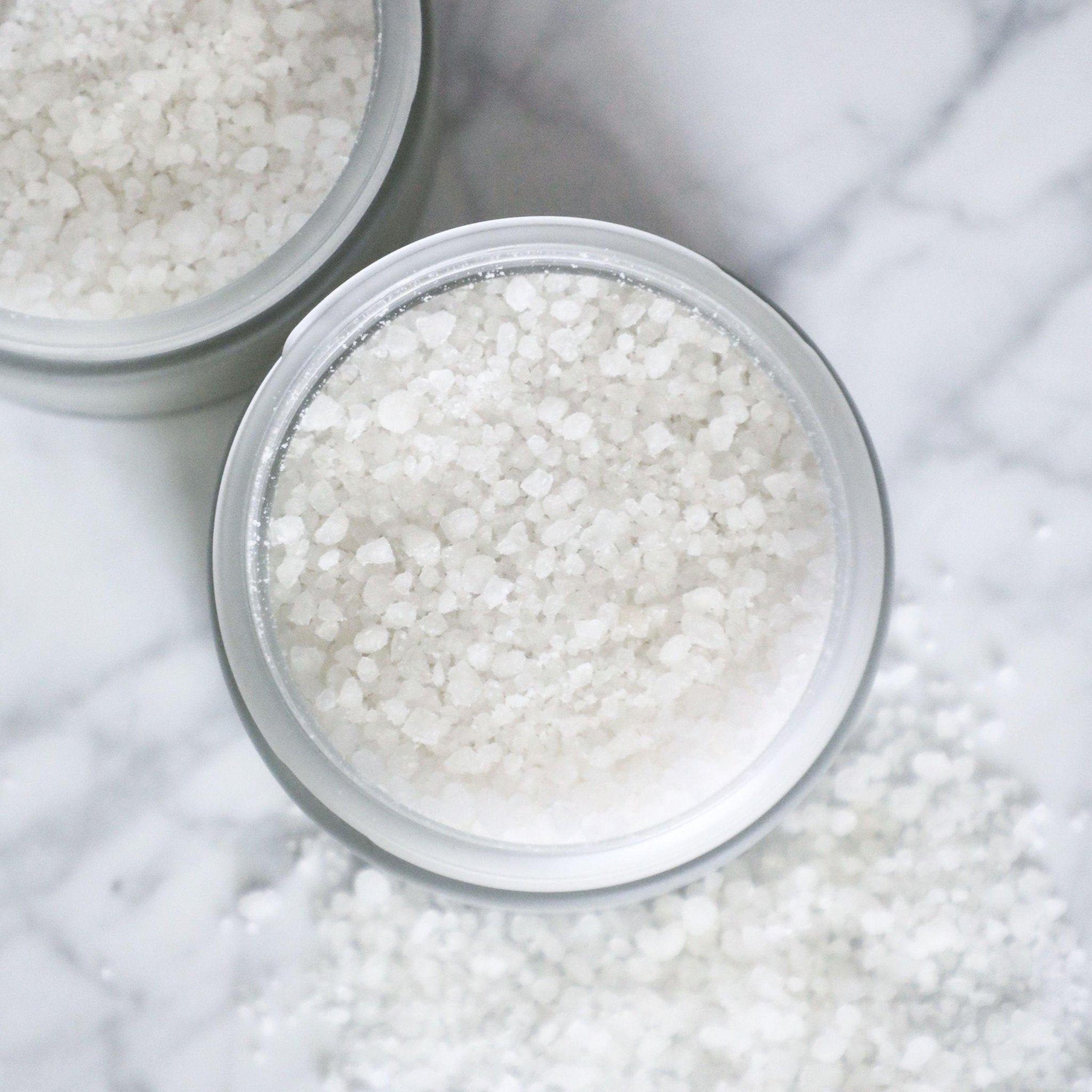 Close-up of two round OLVERUM Bath Salts containers filled with coarse crystals, with mineral soak granules scattered on a white marble surface.