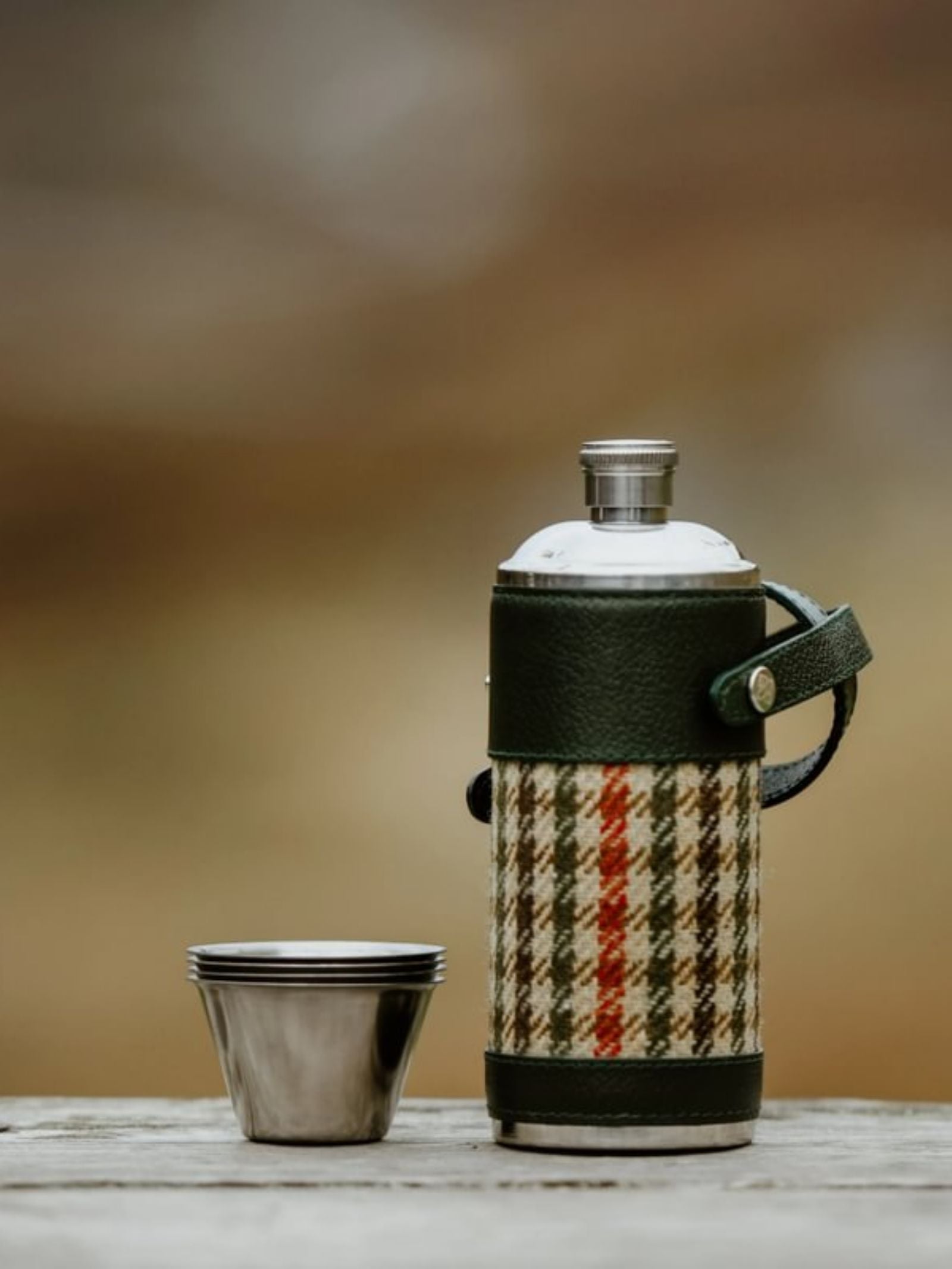 An 8oz Forest Green Hunter Flask by Campbell's of Beauly x Ettinger, wrapped in plaid fabric with green leather accents, sits beside two stacked stainless steel cups on wood, highlighting British craftsmanship against a blurred outdoor backdrop.